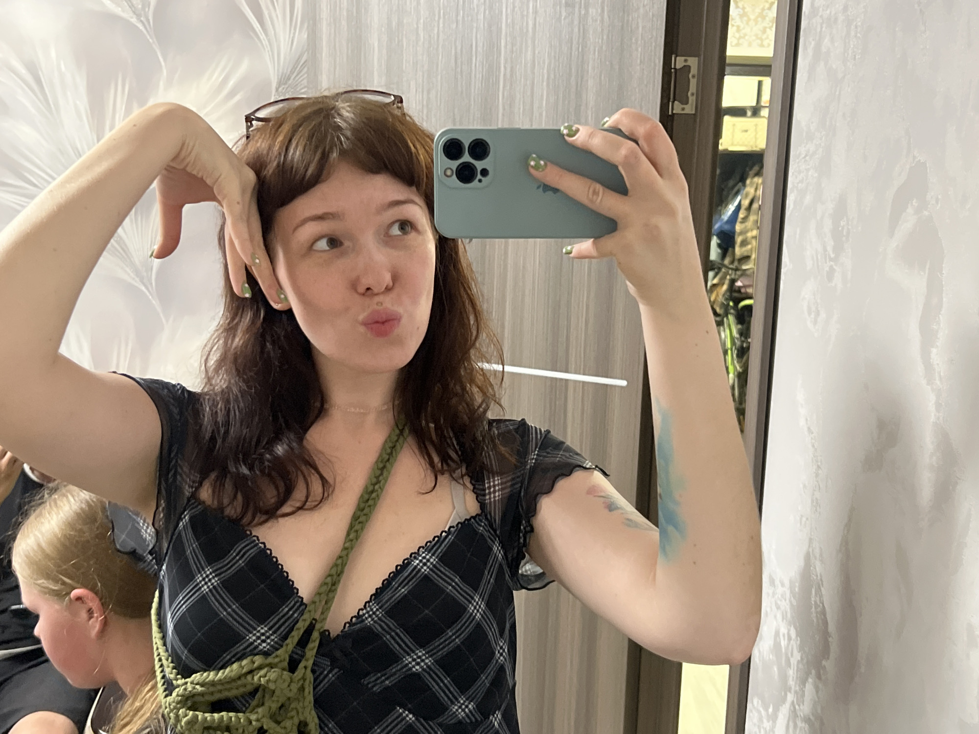 A young woman with brown hair and bangs is taking a mirror selfie in what appears to be a room. She is wearing glasses on top of her head and a black and white plaid dress with sheer sleeves. A green braided strap is draped across her chest. Her left arm is raised, with her hand near her head, and she is puckering her lips. Her fingernails are painted green with white dots. In the background, a younger child with blonde hair is visible from the side, also seated. The wall behind the woman has a textured pattern. Through an open doorway to the right, a glimpse of a bicycle and other items can be seen. The lighting suggests it is daytime.