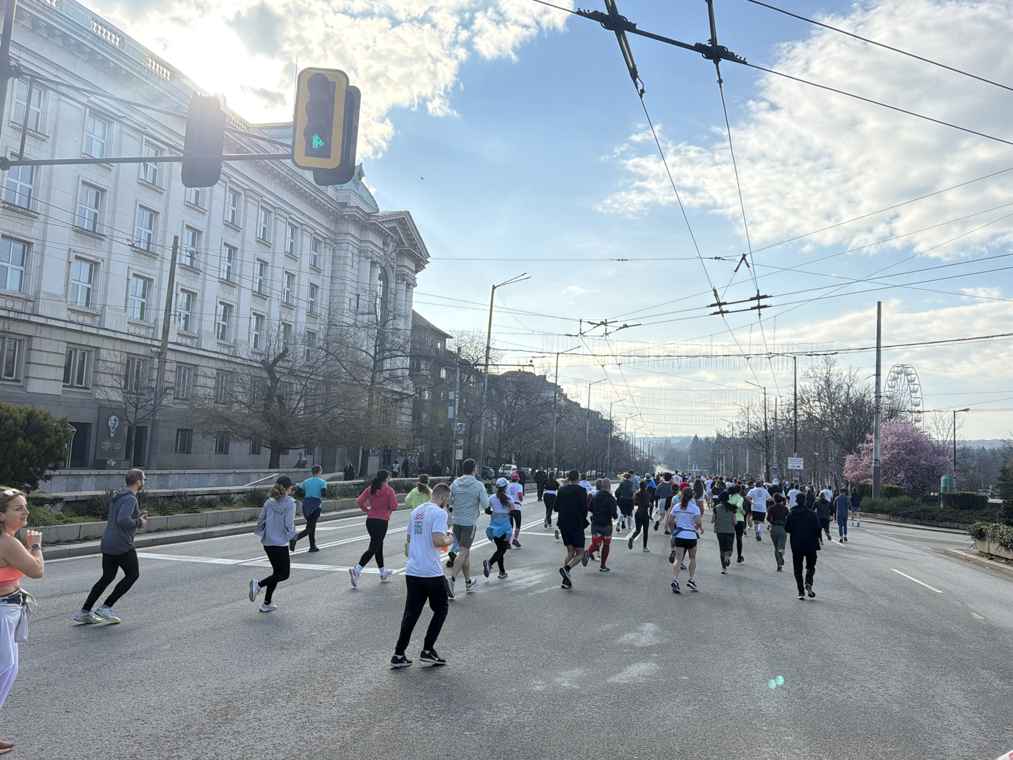 The image depicts a road race in Sofia, Bulgaria. Many runners are seen on a multi-lane road, facing away from the camera. A large, white-stone building is to the left of the road, and a green traffic light is overhead. Overhead electrical lines are strung across the road. In the distance is a Ferris wheel, suggesting a nearby park or entertainment area. Trees line the sides of the road, and some are blooming. The sky is partly cloudy.