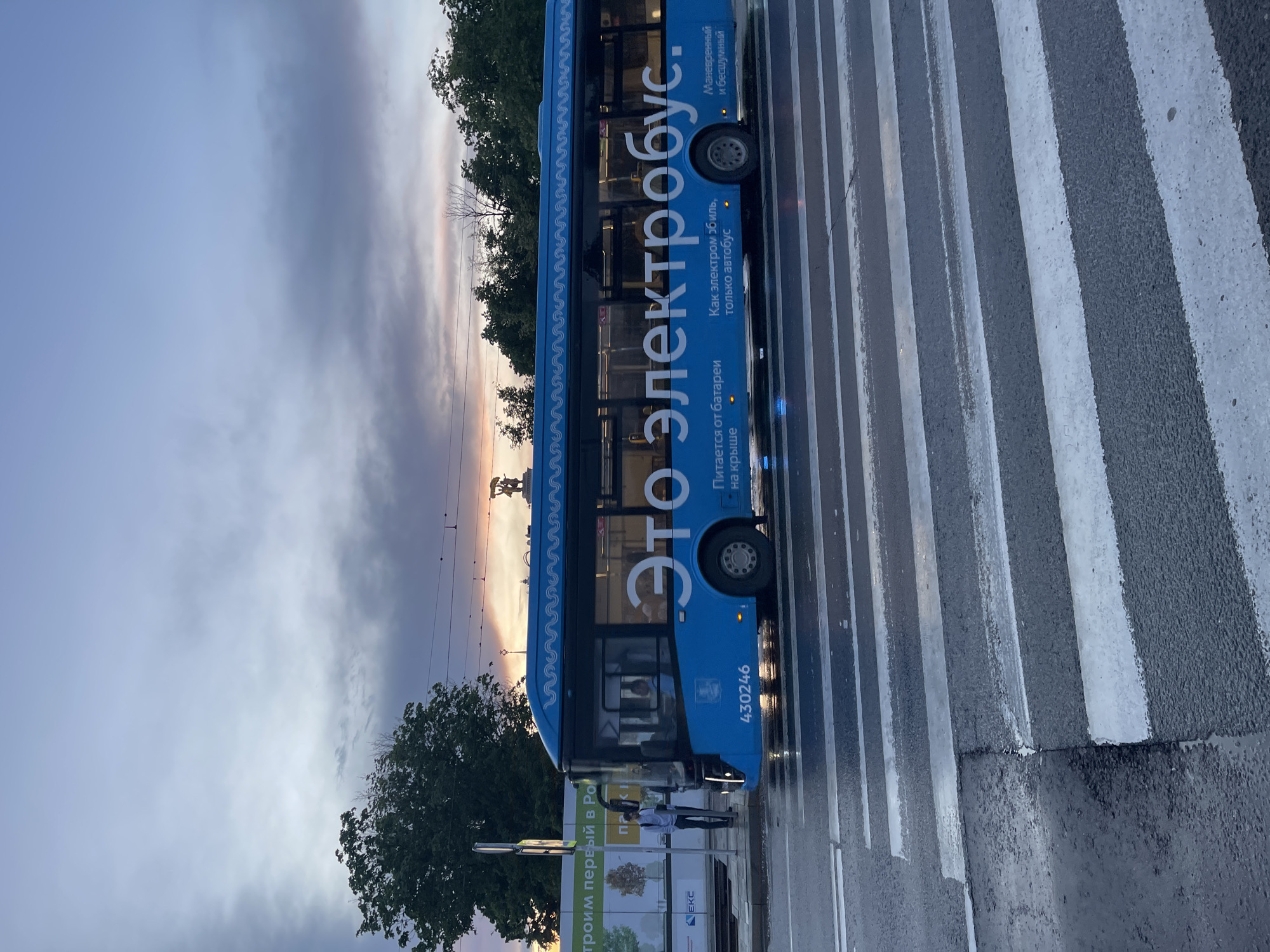 A blue electric bus with the text "Это электробус" ("This is an electric bus") written on it is stopped at a crosswalk. The bus also has text describing it as powered by batteries on the roof and as maneuverable and quiet. Two people are standing near a sign that says "Строим первый в Ро". The background shows trees and a sky with a sunset. The location is in an urban environment with wet roads.