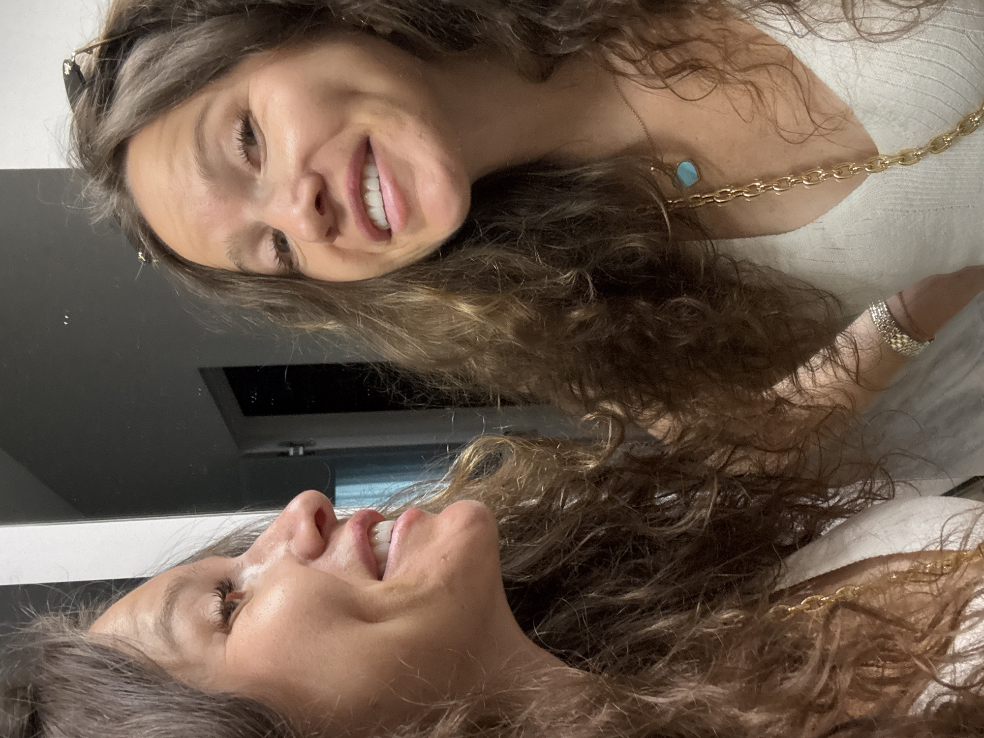 The image shows a close-up of a woman's face and upper body, with a reflection visible. She has long, wavy brown hair and is smiling, revealing her teeth. Her skin appears to have light makeup. She is wearing a white top and a gold necklace with a turquoise pendant, as well as a silver bracelet on her wrist. The background is dark and appears to be a room with a mirror, suggesting the photo was taken in a bathroom or dressing area. There are no visible signs of the city of Unknown, Unknown.