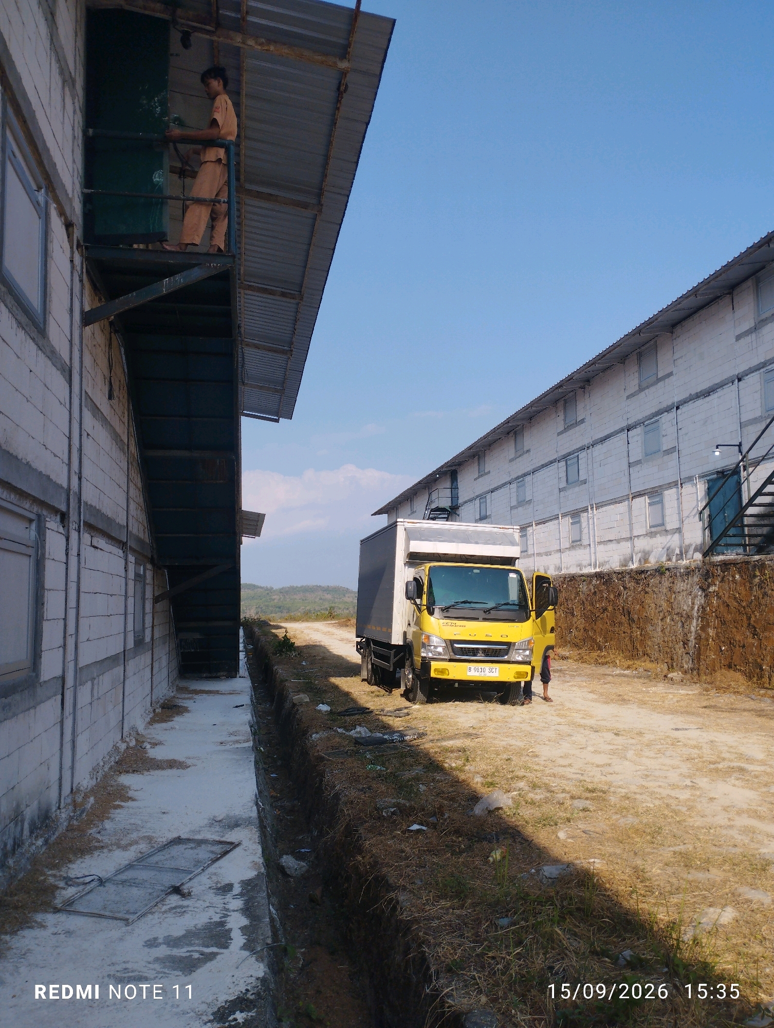 A yellow FUSO truck is parked on a dirt road next to a long, multi-story building constructed of white cinder blocks with a corrugated metal roof. A person wearing a tan uniform stands on a metal walkway attached to the side of the building, holding onto a green door. The building has multiple windows and an external metal staircase leading to the upper floors. The ground in front of the building is a mixture of concrete, dirt, and dry grass, with a trench running along the side. In the distance, rolling hills are visible under a clear blue sky. The image is dated 15/09/2026 15:35.