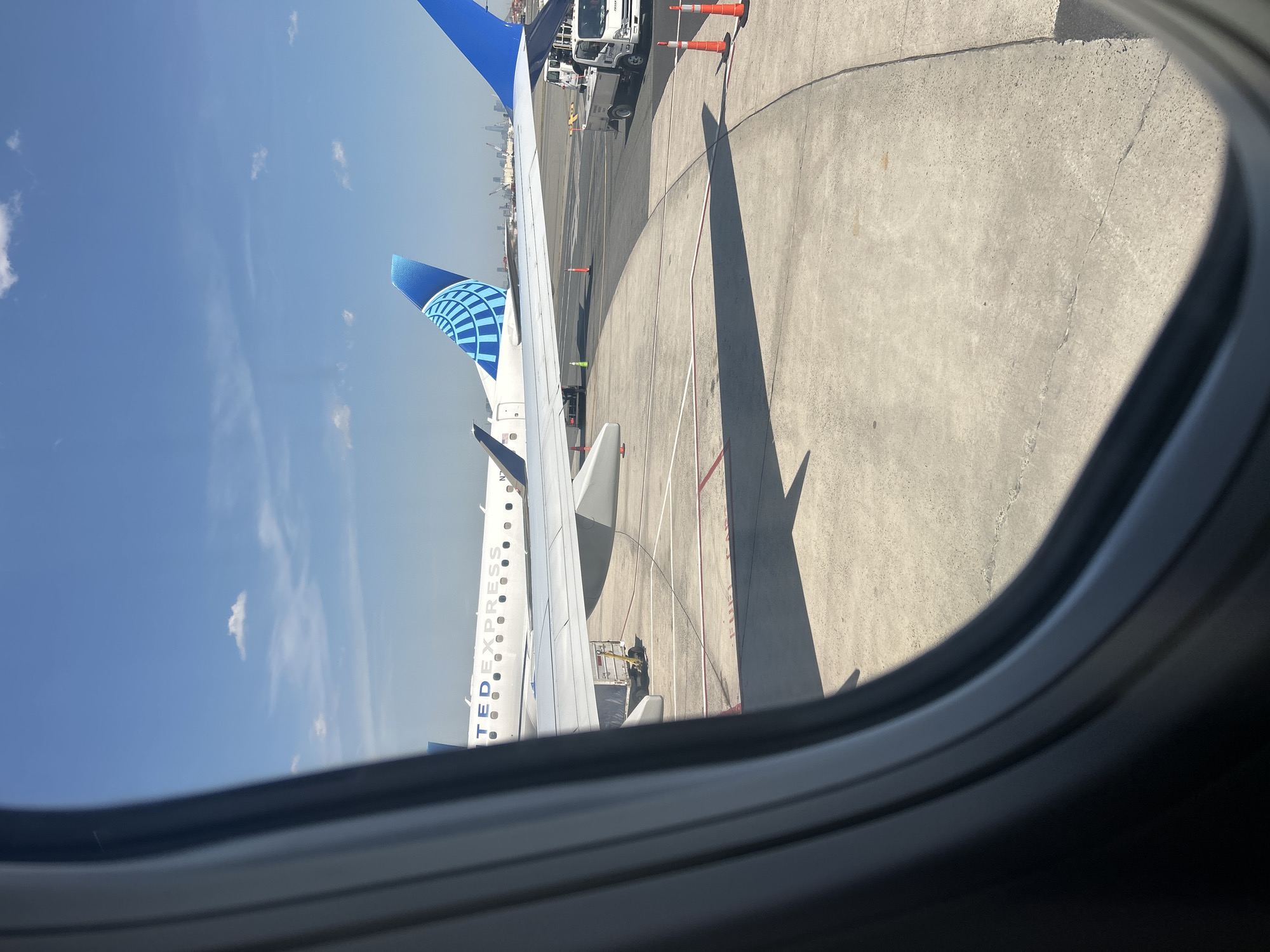 View from inside an airplane window of a United Express Embraer E175 aircraft at Newark Liberty International Airport in Newark, New Jersey. The plane is parked on the tarmac. Ground service equipment is visible around the plane.