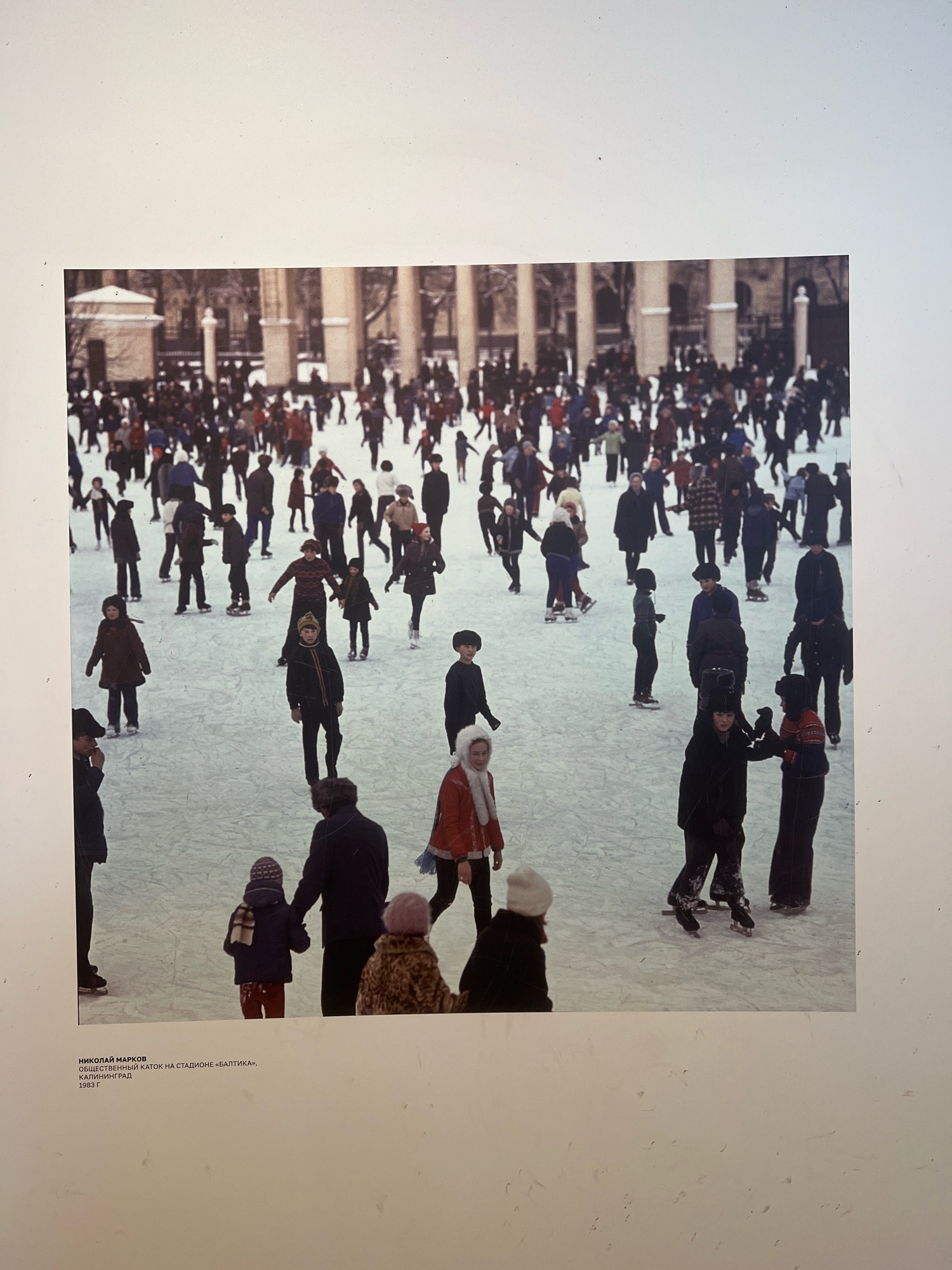 This is a photograph of a crowded outdoor ice rink. Many people, dressed in winter clothing, are ice skating. Some are actively skating, while others appear to be standing or walking on the ice. The background shows large, classical-style columns and buildings, suggesting a public or institutional setting. The text below the image identifies the location as the "Public Skating Rink at 'Baltika' Stadium, Kaliningrad, 1985." The image is a candid shot capturing a moment of leisure and recreation in a winter cityscape.