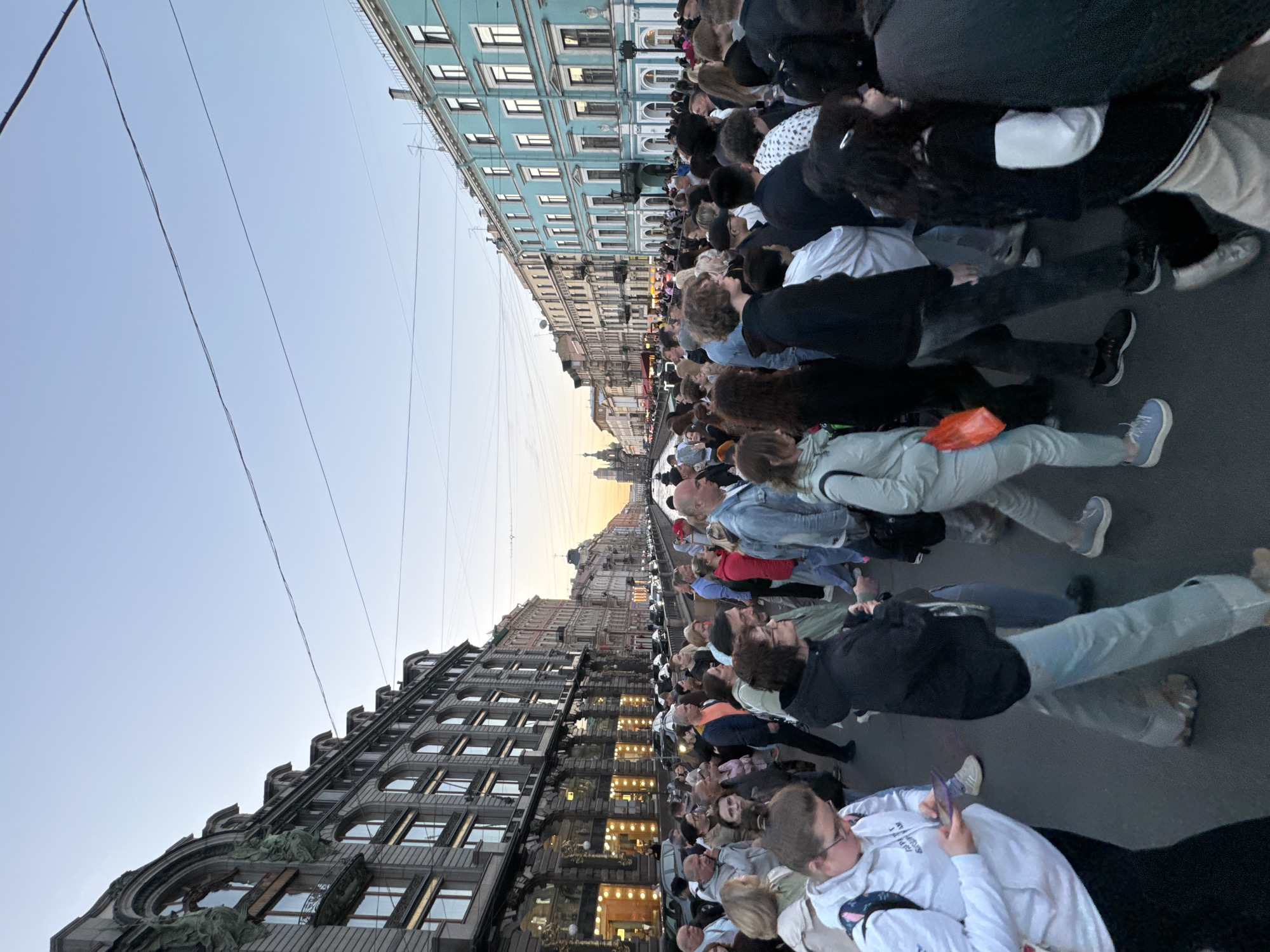 The image captures a crowd of people walking along a street in Saint Petersburg, Russia, likely during the evening given the soft light. The architecture suggests a historic European city, with grand, multi-story buildings lining both sides of the street. The building on the left appears to be made of stone with arched windows and ornate detailing. The building on the right is painted a distinct teal color and features a similar architectural style. A canal or river is visible in the background, spanned by a bridge, and more buildings stretch into the distance, culminating in church spires under a pale sky. People are dressed in casual, contemporary clothing, and many are walking in the same direction, suggesting a pedestrian thoroughfare or event. Some individuals are looking at their phones, while others are engaged in conversation. The overall atmosphere appears to be one of a bustling urban environment.