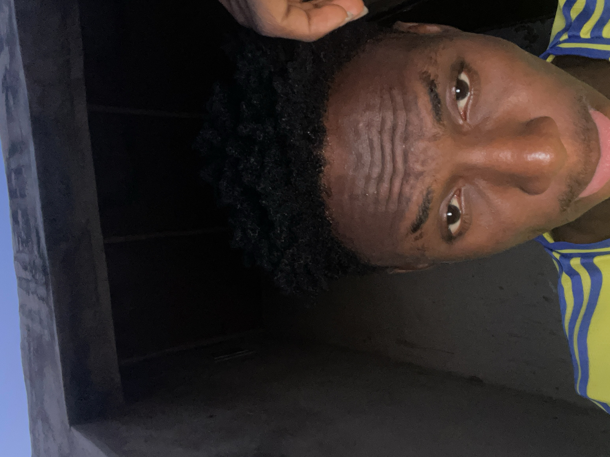 A close-up, slightly tilted image shows a dark-skinned person with curly black hair. The person is wearing a yellow and blue striped shirt. The background appears to be an unlit, concrete structure. It is impossible to determine a specific location in the city of Lokoja, Nigeria from the image.