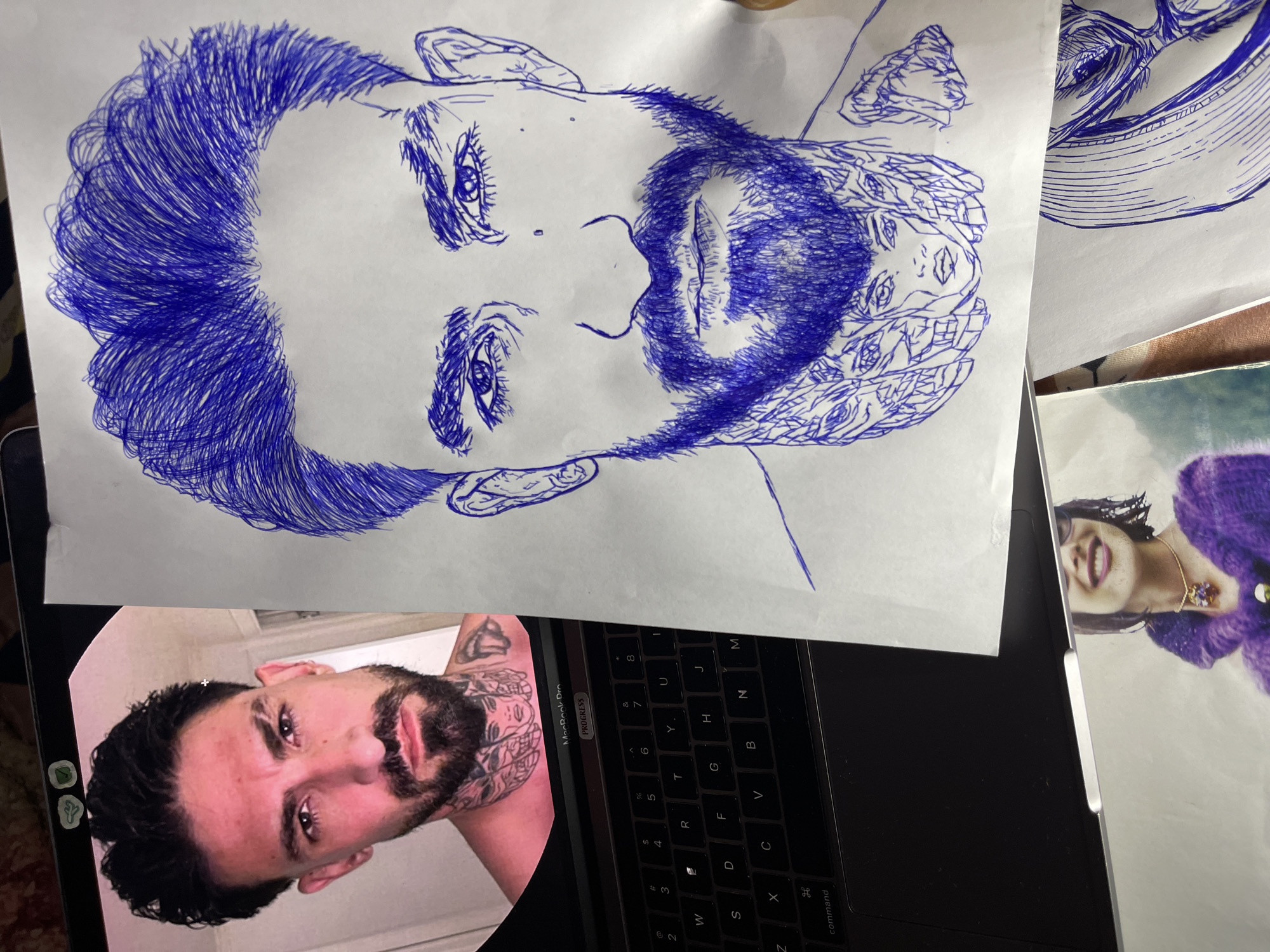 The image displays a close-up view of a person's face, captured from the chest up. The subject has dark hair, a beard, and tattoos on their neck and chest. The photograph appears to be taken from a laptop screen, which is visible in the lower portion of the image, displaying a keyboard and trackpad. To the right of the laptop screen, a blue ink drawing is laid out on a white surface, depicting a detailed portrait of the same individual. The drawing includes intricate details of the hair, eyes, beard, and neck tattoos, which feature multiple faces. In the foreground, another photograph is partially visible, showing a person wearing a purple sweater and a necklace. The overall scene suggests an artistic process, possibly the creation of a portrait from a reference image. The image does not contain any information that would allow for the determination of its location in the city of Jos, Nigeria.