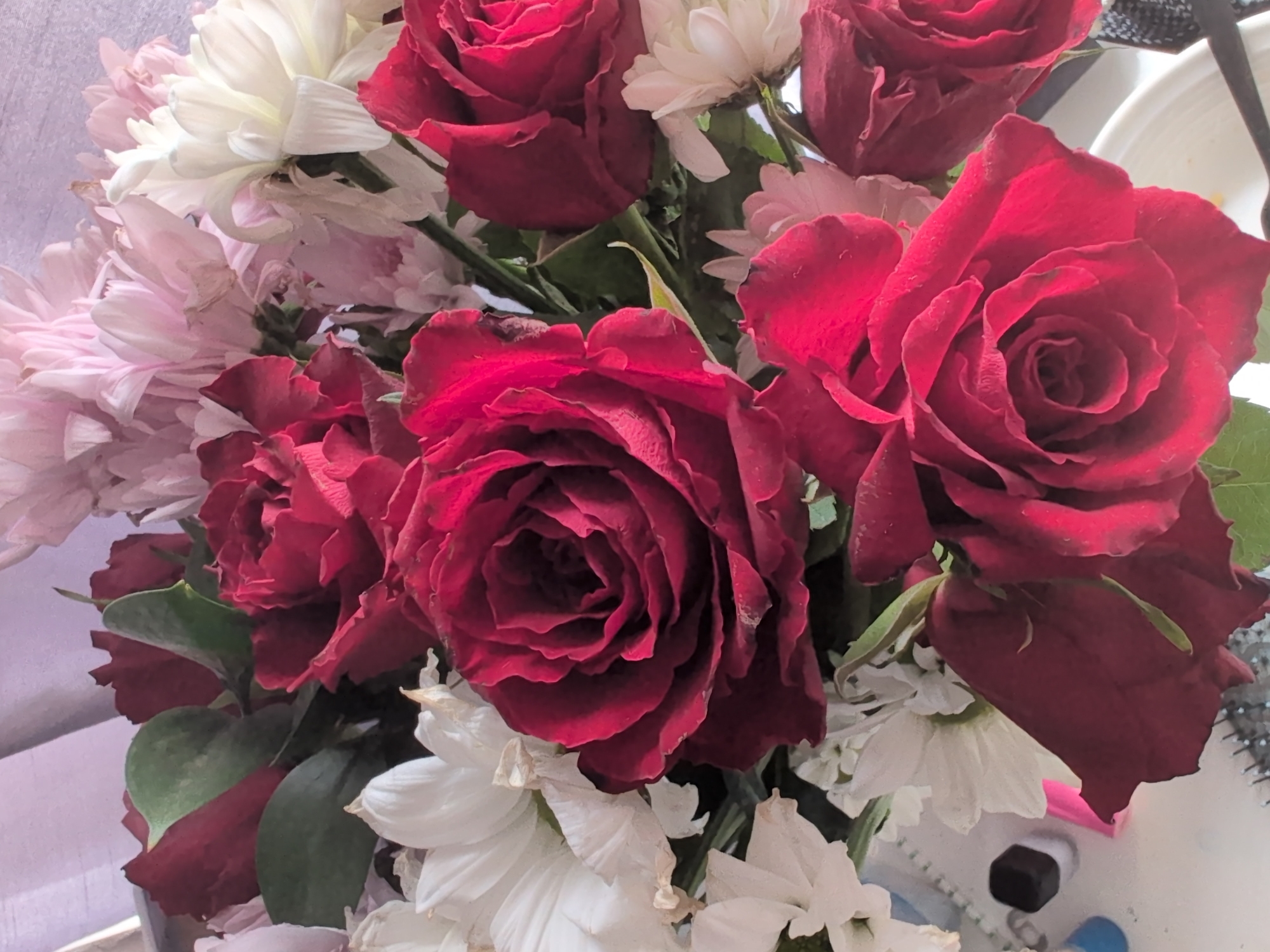 The image shows a close-up of a bouquet of flowers, featuring predominantly deep red roses interspersed with white and pale pink chrysanthemums. The flowers are arranged in a dense cluster, with some green leaves visible. The background is softly lit and out of focus, suggesting an indoor setting. There are no people or discernible city elements visible in the image, making it impossible to determine a specific location within Oxford, United Kingdom. The scene is static, with the focus entirely on the floral arrangement.