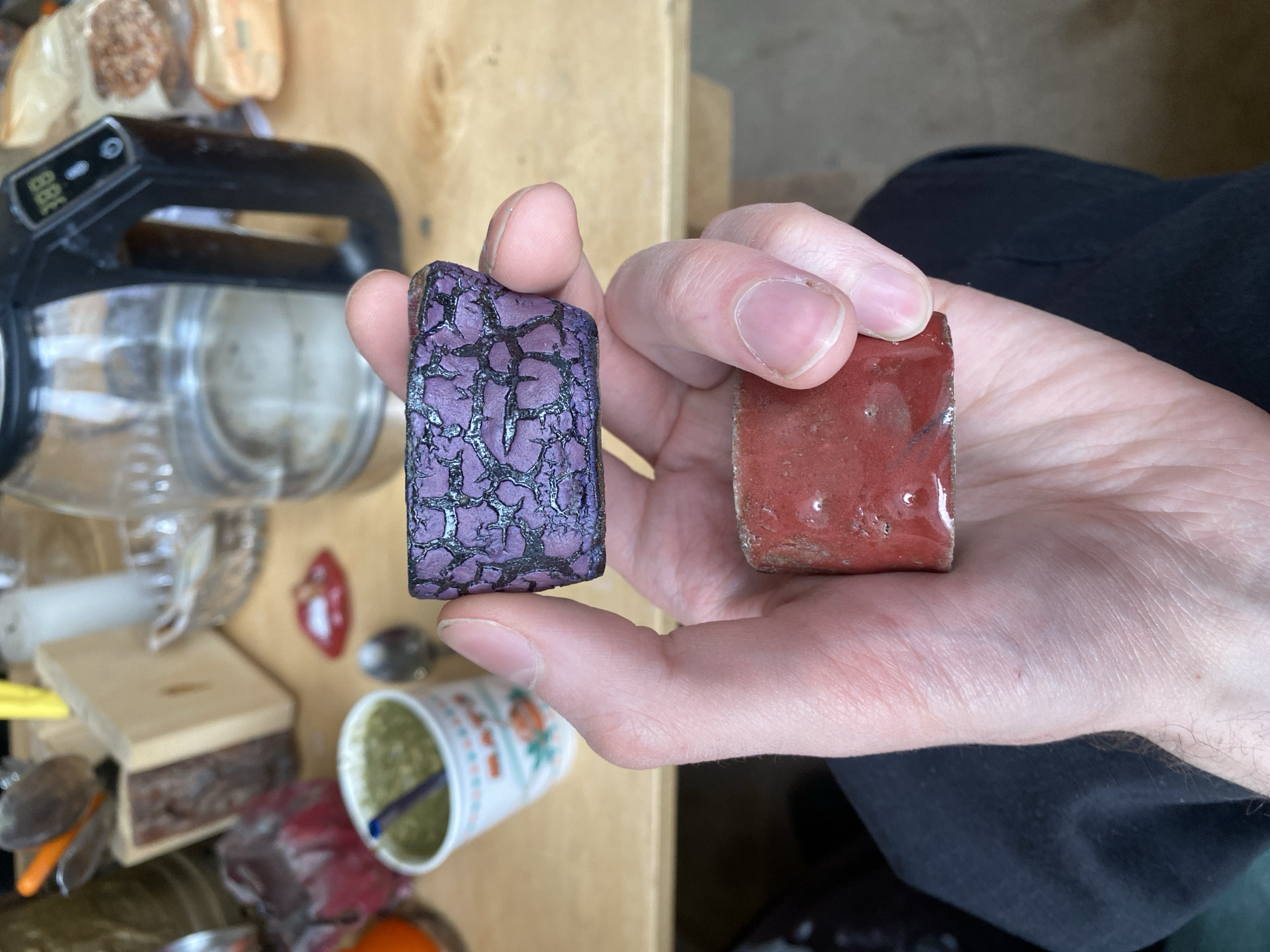 A photograph shows a fair-skinned hand holding two small, rectangular objects against a cluttered background. The object in the left hand is purple with a black crackle pattern, appearing somewhat glossy. The object in the right hand is red and also appears to be coated with a glossy substance. The hand holding the objects is positioned against a backdrop of items on a wooden surface, including a clear plastic container with a black handle, a small wooden box, utensils, a cup with green contents, and other miscellaneous objects. The overall scene is brightly lit, and no individuals are visible except for the hand and a portion of a dark garment near the bottom right corner of the frame. There is no evidence to indicate the location of the city of Moscow, Russia.