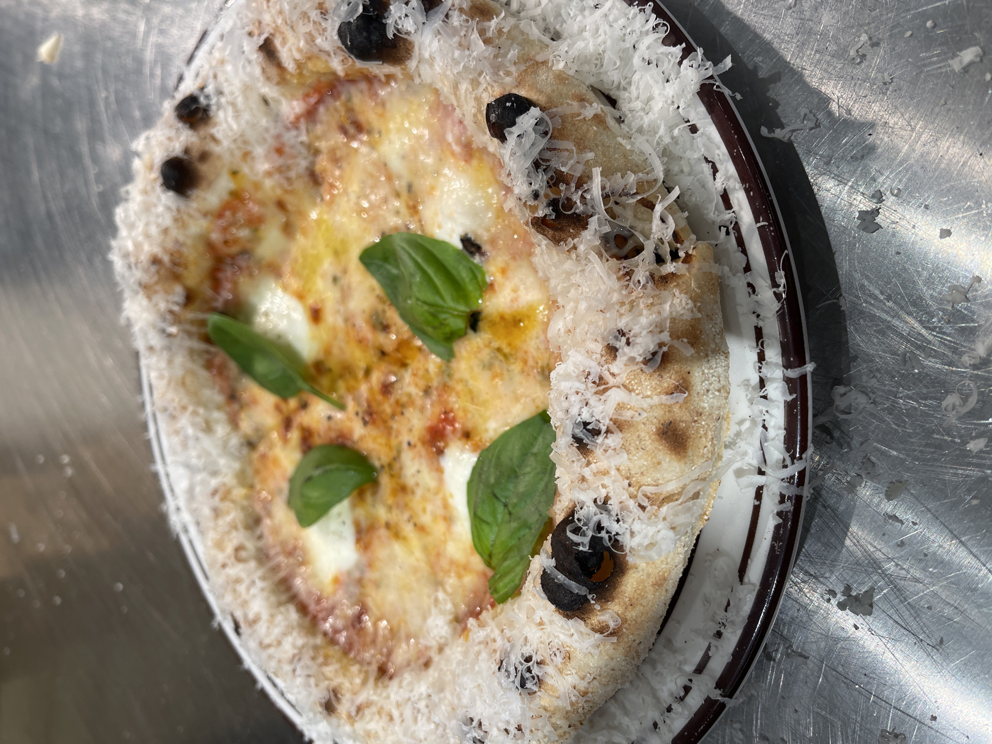 A slightly high-angle, medium shot shows a pizza on a plate sitting on a stainless-steel surface. The pizza has a thin crust that is charred in places. The toppings include cheese, tomato sauce, basil leaves, and what appear to be black olives. The crust is heavily covered with grated cheese. The plate is white with a dark brown or red rim. The stainless-steel surface has visible scratches. The location is unspecified, but the query mentions Belgrade, Serbia. No people are visible.