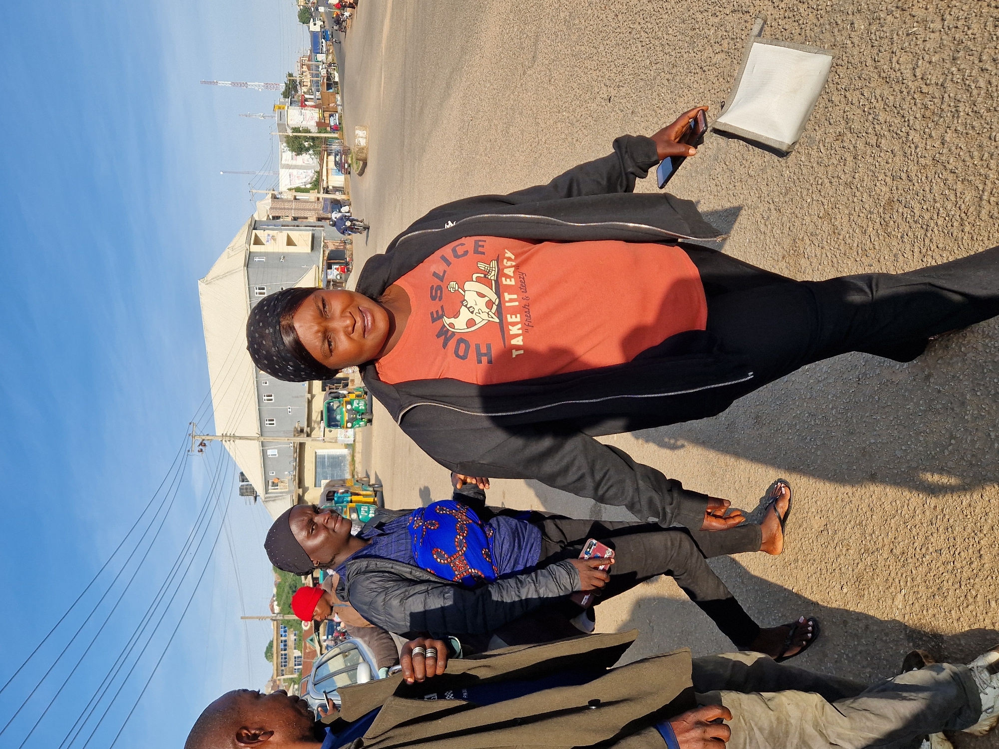 A medium shot shows three people walking on a paved road in Kabong, Nigeria. The foreground features a woman in the center facing the camera, wearing a black headscarf, a black zip-up jacket over an orange t-shirt with "HOME SLICE TAKE IT EASY" printed on it, and black pants. She is holding a mobile phone and a small, light-colored pouch in her right hand. To her left, a person wearing a dark jacket and a dark headscarf is partially visible, also holding a mobile phone. To the left of the central woman, a person in a brown jacket and light-colored pants is seen walking away from the camera, with their left arm extended. In the background, a street lined with buildings and power lines is visible under a clear blue sky. Several tricycles and vehicles are present on the road. The sun casts long shadows on the ground.