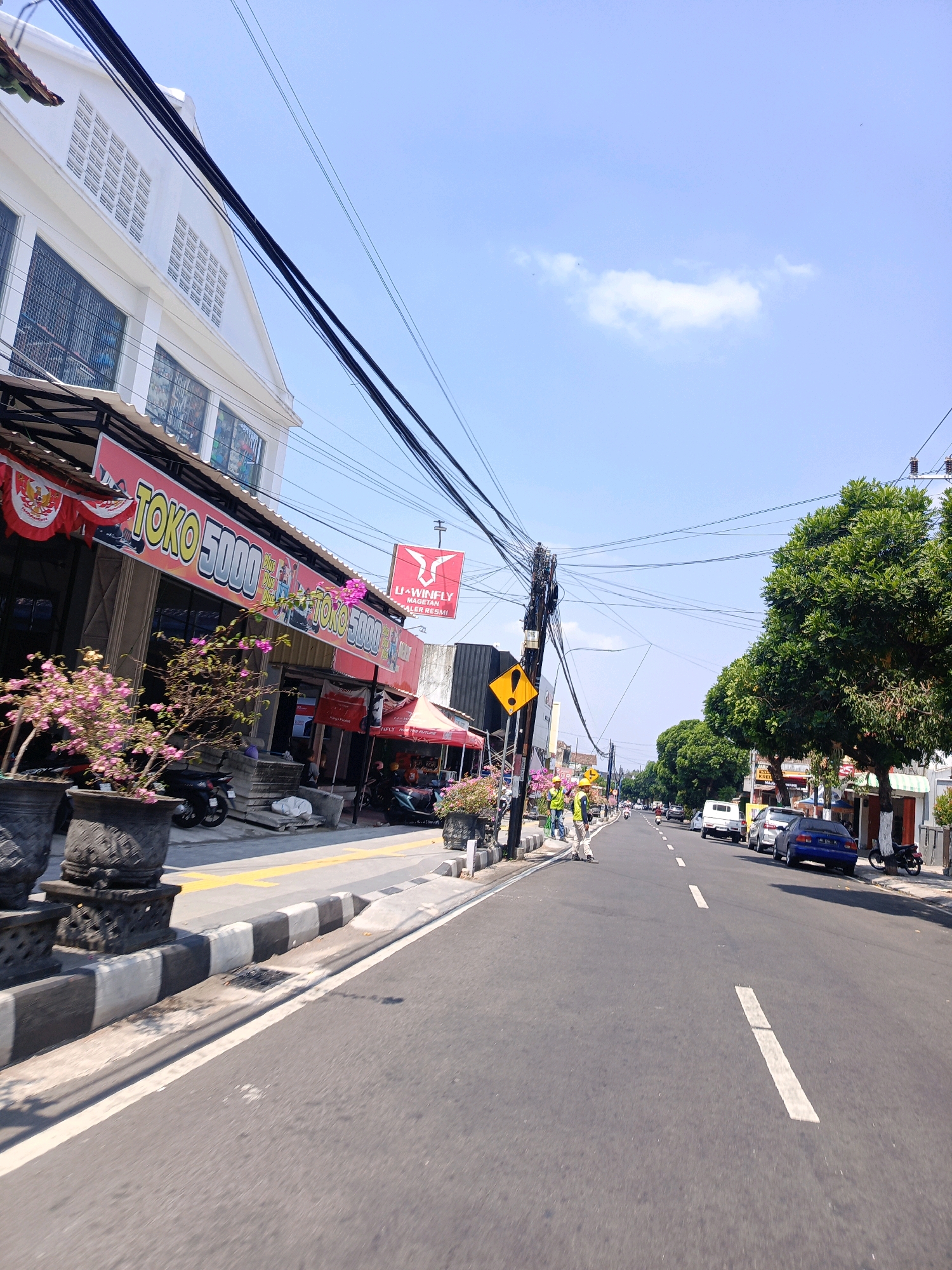 The image depicts a street scene in the city of Magetan, Indonesia, on a clear, sunny day. The street is paved with asphalt and has visible white lane markings. On the left side of the street, there is a building with a white facade and several windows. A prominent red sign with white lettering reads "TOKO 5000". In front of the building, there are large potted plants with pink flowers and motorcycles parked. A few people, some wearing yellow vests, are visible on the sidewalk near a utility pole cluttered with numerous wires. A yellow warning sign with a black exclamation mark is attached to the utility pole. Further down the street, there are more buildings, trees, and several parked cars, including a blue sedan and a white van. The sky is bright blue with a few wispy clouds.