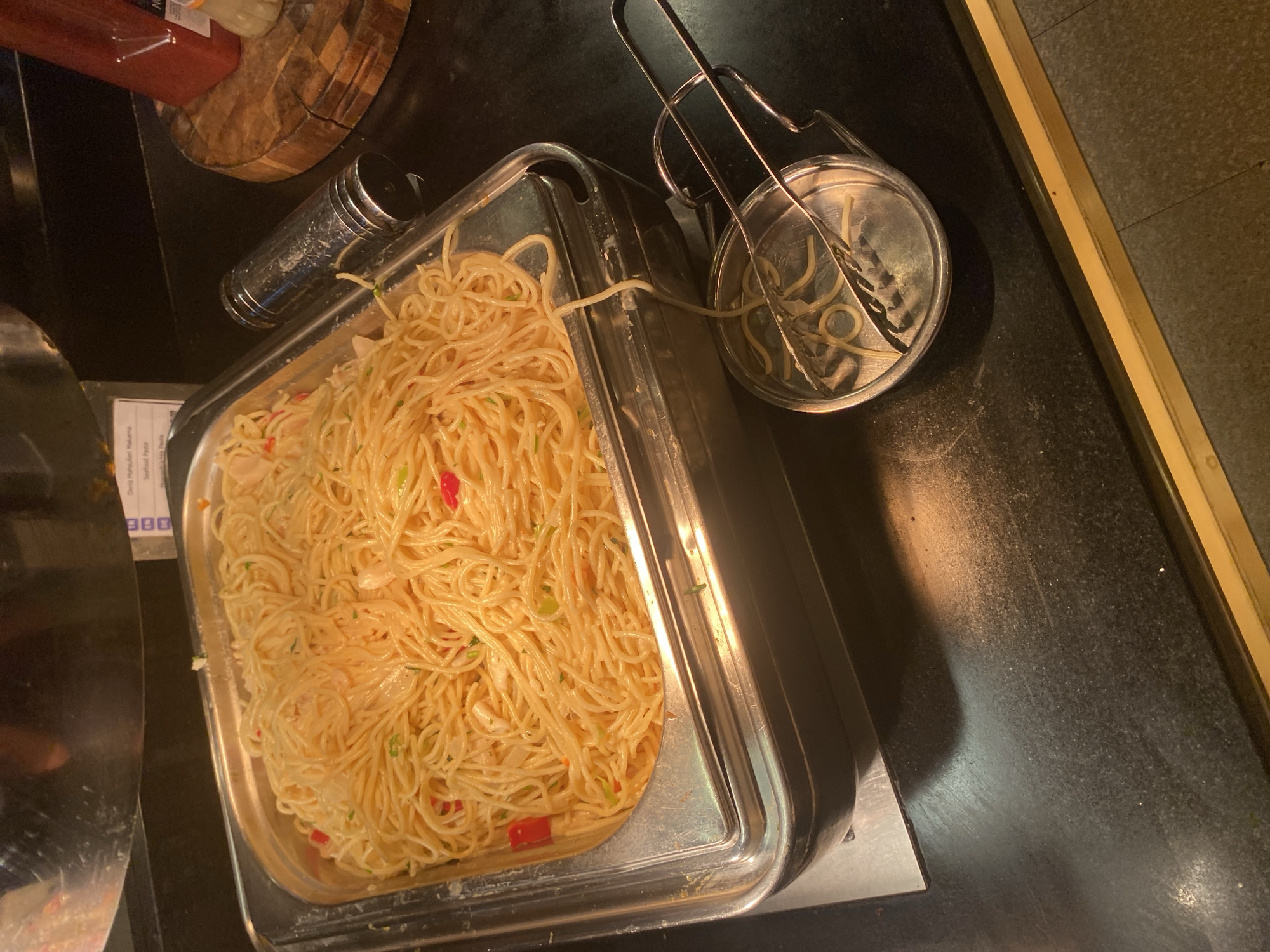 A close-up, high-angle shot shows a metal serving tray filled with spaghetti in a buffet setting. The spaghetti appears to be tossed with a light sauce and contains small pieces of red pepper and green herbs. A metal tong is resting on a small metal dish next to the serving tray, with a few strands of spaghetti on it. In the background, a sign with text in multiple languages, including "Deniz Mahsulleri Hakama" (likely Turkish for "Seafood Hakama") and "Seafood Pasta," is partially visible. To the right, a wooden cutting board with a red bottle and a small white jar is visible. The serving tray and surrounding counter are made of metal and a dark, textured surface, respectively. The lighting suggests an indoor environment, possibly a restaurant or hotel buffet. The specific location in Aksu, Turkey, cannot be determined from the image.