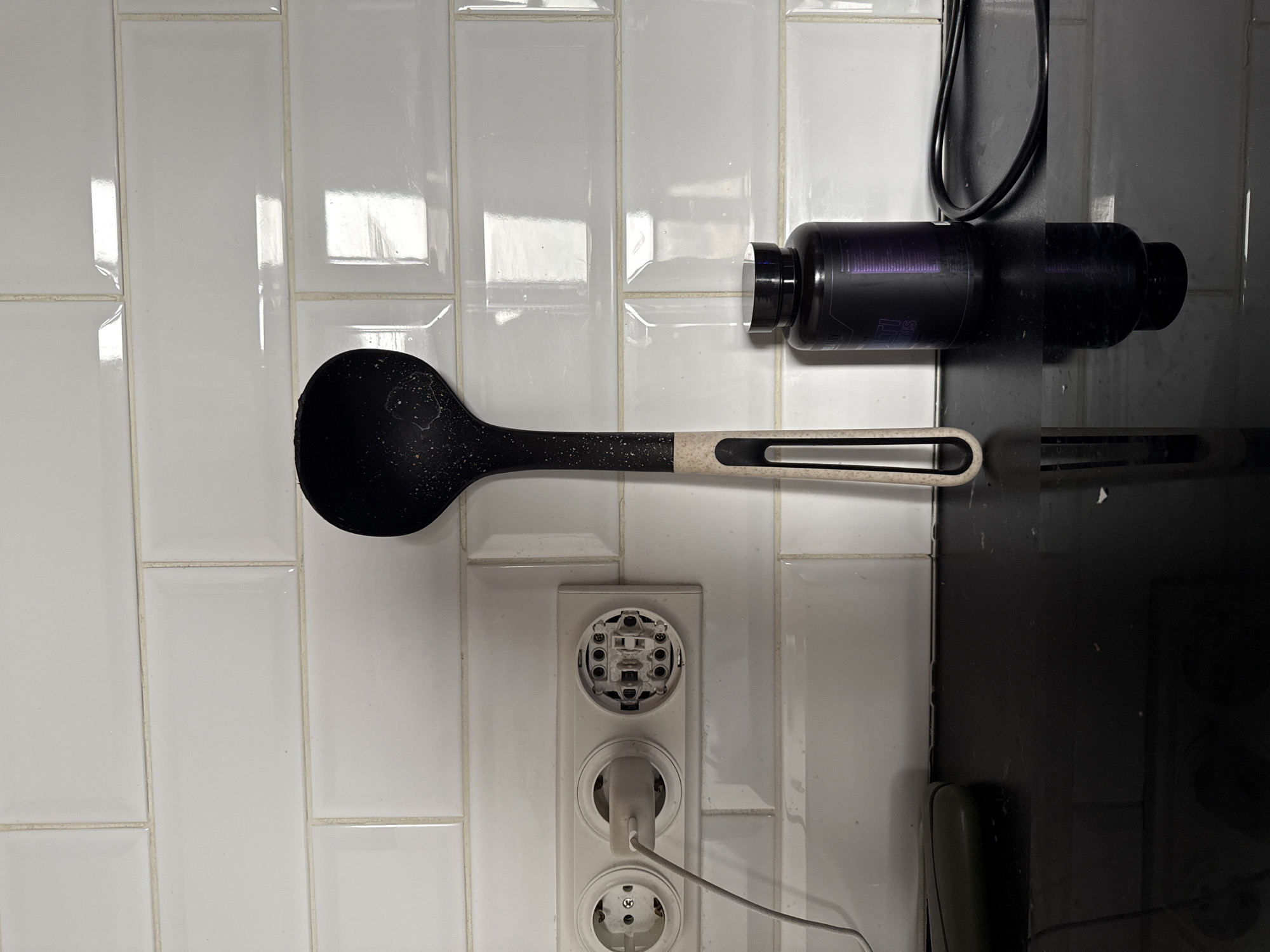 A stark, clinical view of a kitchen environment is presented. The backdrop is a white tile wall, reflecting light. A black ladle rests against the wall above an electrical outlet. The outlet consists of three receptacles, the top one missing its cover, exposing its internal components. A plug is partially inserted into the middle receptacle, a gray cable extending from it. A black bottle with a cable running up is placed on the adjacent surface. The surface to the right of the wall appears to be a black countertop, showing signs of use, with a hint of dark green objects in the lower right corner. This image could potentially be located in the city of Vidnoye, Russia.
