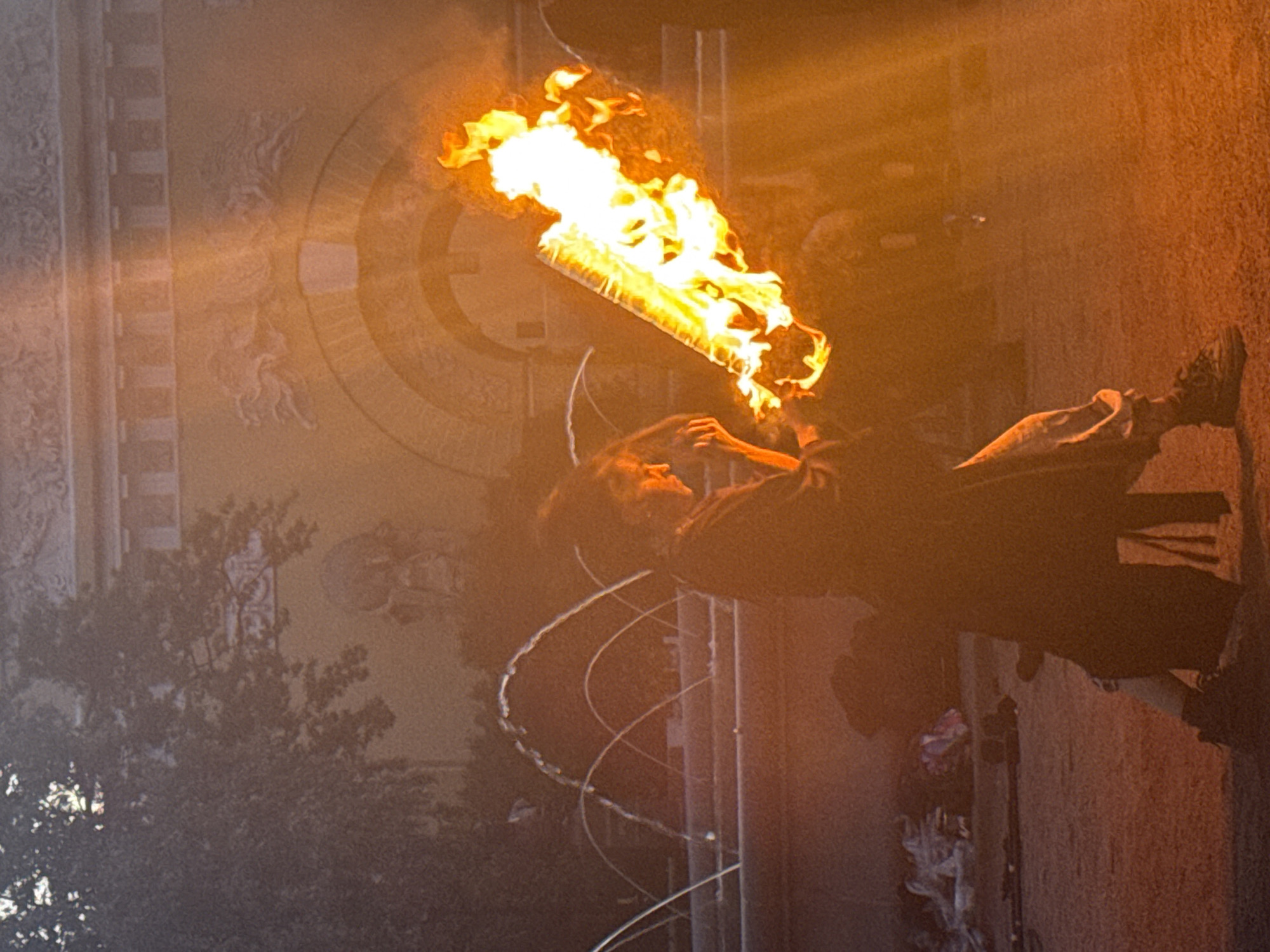 The image depicts a street performer in Saint Petersburg, Russia, engaging in a fire-spinning act in front of an ornate building. The performer, dressed in dark clothing and light-colored pants, is holding a flaming object that appears to be a staff or a torch, with bright flames extending upwards. The background features a building with classical architectural elements, including arches and decorative reliefs, possibly part of the historic city. Several arcs of water from a fountain are visible in the mid-ground, suggesting the performance is taking place in a public square or park. The lighting is dramatic, with the fire illuminating the scene and casting a warm glow. The overall impression is one of a dynamic and visually striking street performance in an urban setting.