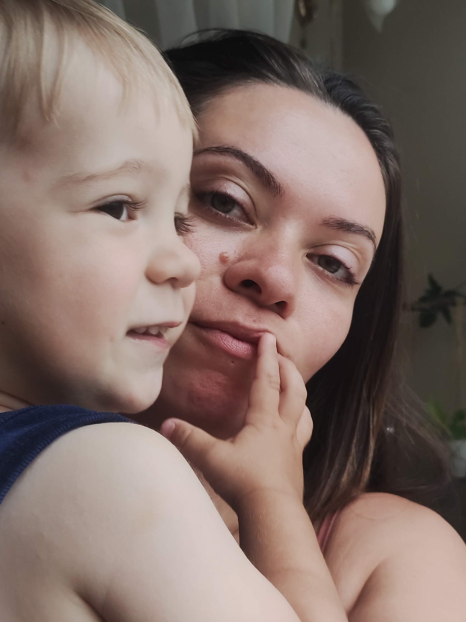 An over-the-shoulder, selfie-style, medium shot captures a woman and a young child in close proximity. The woman, in the foreground and on the right, has dark brown hair and is looking towards the camera with a thoughtful expression, her finger touching her lower lip. She is wearing a light pink top. The child, positioned on the left and in profile, has light blonde hair and is looking off to the left, with a hint of a smile. The child is wearing a dark blue top. The background is softly blurred, suggesting an indoor setting with a window and some greenery visible. The image appears to have been taken in Chaikovsky, Russia.