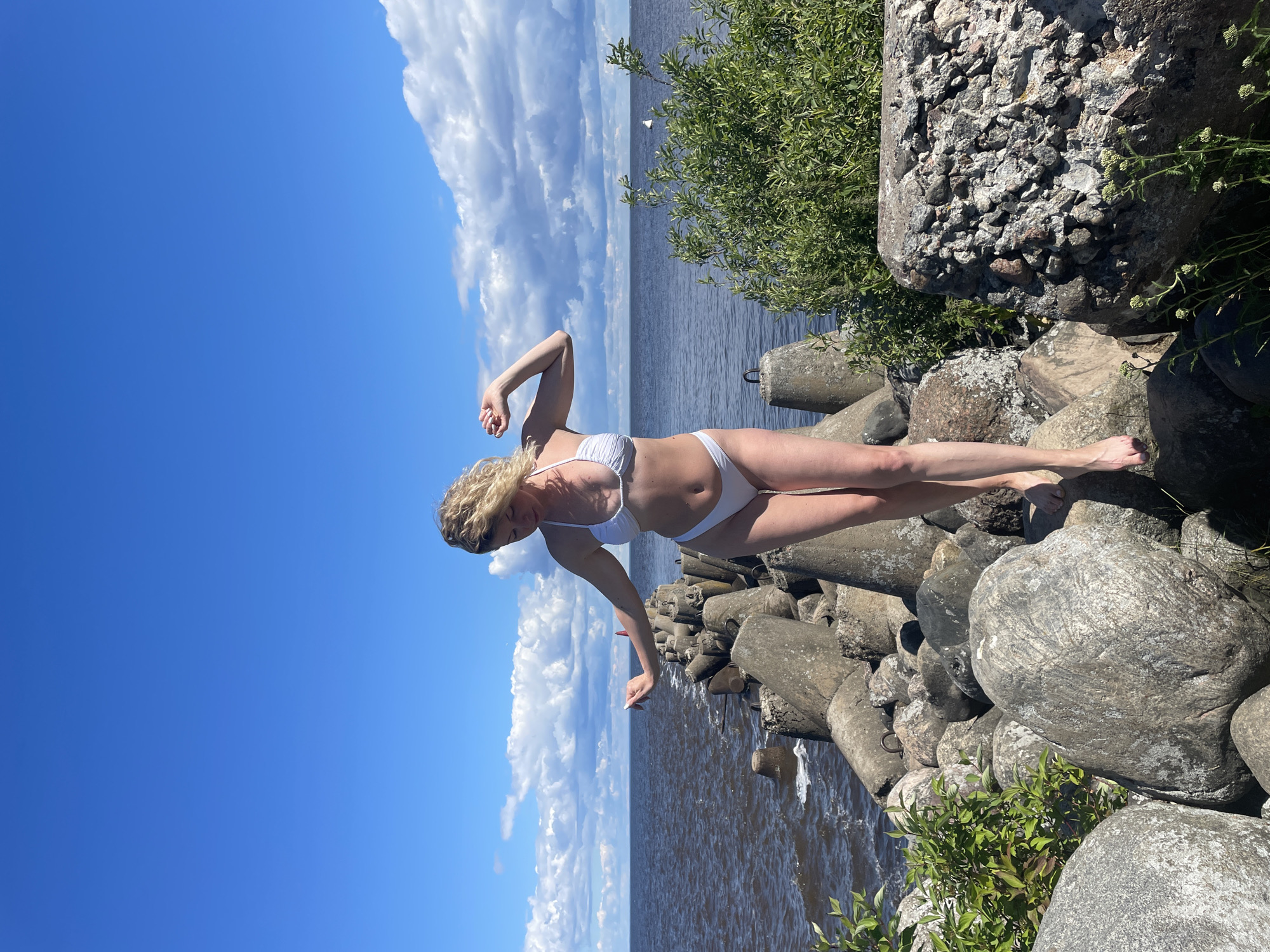 A blonde woman in a white bikini stands on a rocky breakwater under a blue sky with scattered clouds. She is facing slightly away from the camera, with her head tilted down and her arms outstretched, one raised and bent at the elbow, the other extended to her side and slightly bent. The breakwater is constructed of large, rough-hewn concrete blocks and rocks. To the left, the dark water of a lake or sea is visible, with waves lapping at the breakwater. To the right, lush green foliage grows among the rocks. The scene appears to be located on the shore of Lake Beloye in Belozersk, Russia.