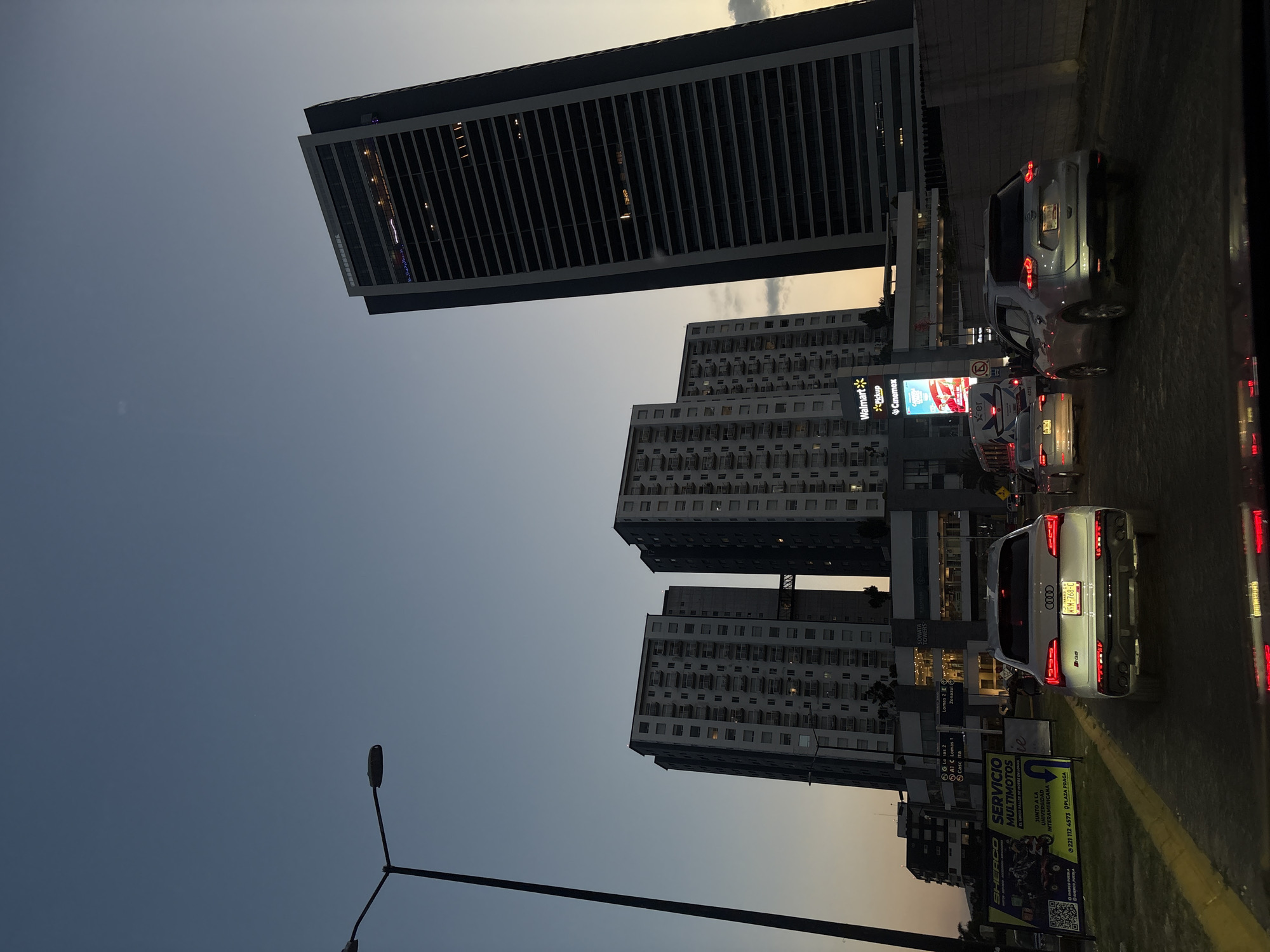 The original image is rotated 90 degrees counter-clockwise, depicting an urban street scene at dusk or early evening in San Bernardino Tlaxcalancingo, Mexico. When viewed correctly, the perspective is from ground level, looking up towards a street lined with buildings and vehicles.
The sky transitions from a light blue-grey at the top to a warmer orange-yellow closer to the horizon, indicating the sun has recently set or is about to set. Artificial lights are visible on buildings, street signs, and the taillights of vehicles, illuminating the scene.
Dominating the background is a tall, modern skyscraper with a dark facade and numerous horizontal elements. Below and to its right, several multi-story apartment buildings with repetitive window patterns are visible. One building prominently features a lit sign displaying "Walmart Pickup" and "Cinemex" logos.
In the midground and foreground, a street is occupied by several cars, suggesting traffic. A silver Audi SUV with illuminated taillights, bearing the license plate "WRM-768-C," is a prominent vehicle. Another white vehicle with "Ker" visible on its side is also present in the traffic.
Various signs are scattered along the roadside. A street sign lists "SONATA TOWERS" and directional information such as "Lomas 2," "Lomas 1," and "Casc Ita." A larger billboard advertises "SERVICIO MULTIMOTOS," mentioning "JUNTO A LA UNIVERSIDAD INTERAMERICANA," "PLAZA PRAGA," and a phone number: "221 112 4573." A black street light pole with two lamps is visible in the lower left foreground. The scene captures a typical bustling city environment during the transition from day to night.