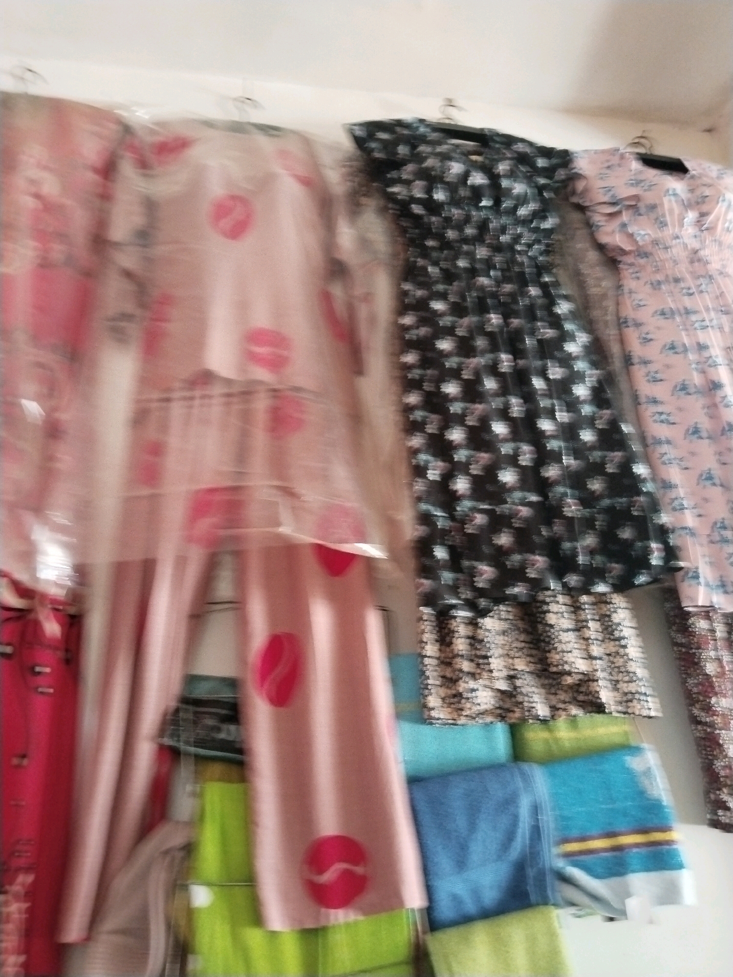 The image shows several pieces of clothing hanging on a rack against a white wall. The clothing includes a pink pajama set with a kiss mark pattern, a black dress with a floral pattern, and a pink dress with a floral pattern. Below the hanging clothes, there are several folded towels in various colors, including green, blue, and turquoise. The lighting is somewhat dim, and the image is slightly blurred, making it difficult to discern fine details of the clothing and the environment. The scene appears to be inside a shop or a home. The city of Prigi, Indonesia, is not identifiable from the visual information present in the image.