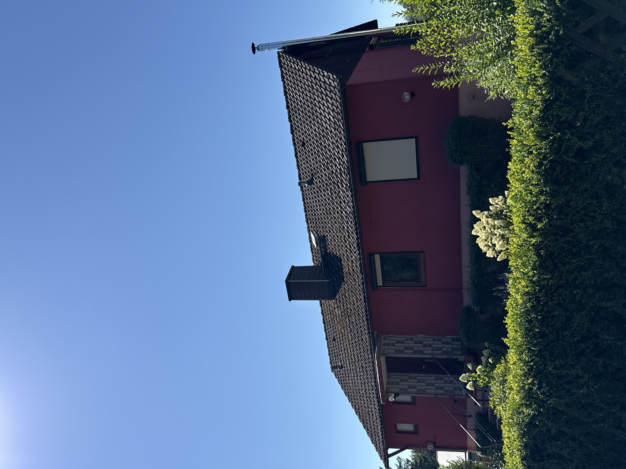 A red, single-story house with a dark tiled roof stands under a clear blue sky. The house has a prominent dark brick chimney on the roof and a silver metal chimney extending from the side of the roof. There is a single door with a small covered porch and stairs leading up to it. Several windows are visible on the house, including one with a rolled-down shutter. Lush green bushes and manicured hedges surround the house. The image was taken on the property of a residence in Bad Sachsa, Germany.