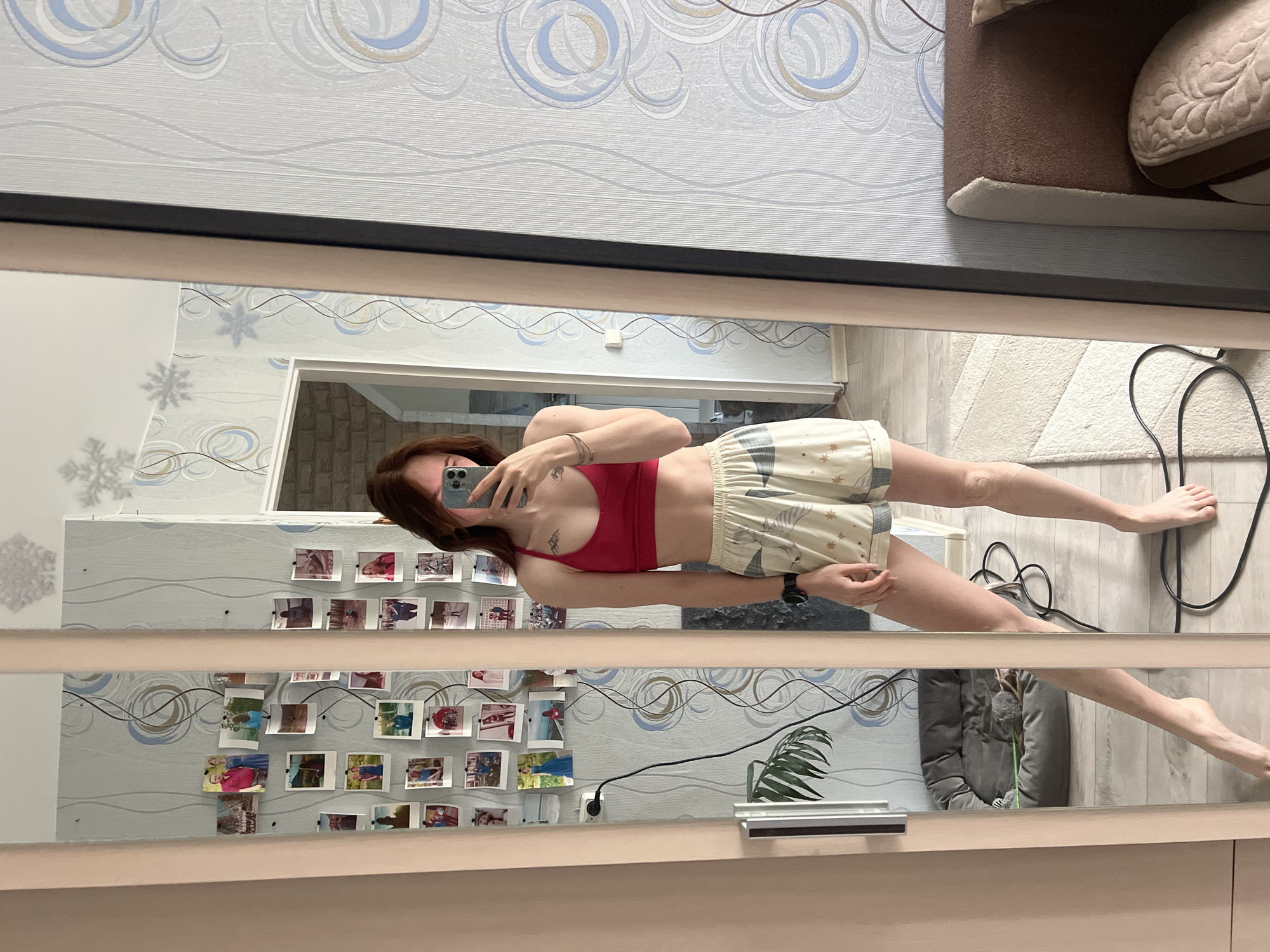 A young woman is taking a selfie in a mirror. She is wearing a red sports bra and light-colored shorts. Her hair is brown and pulled back. She is holding a cell phone in her right hand. The room in the background has patterned wallpaper and a collection of photographs on the wall. The location may be Chaikovsky, Russia, based on prior knowledge.