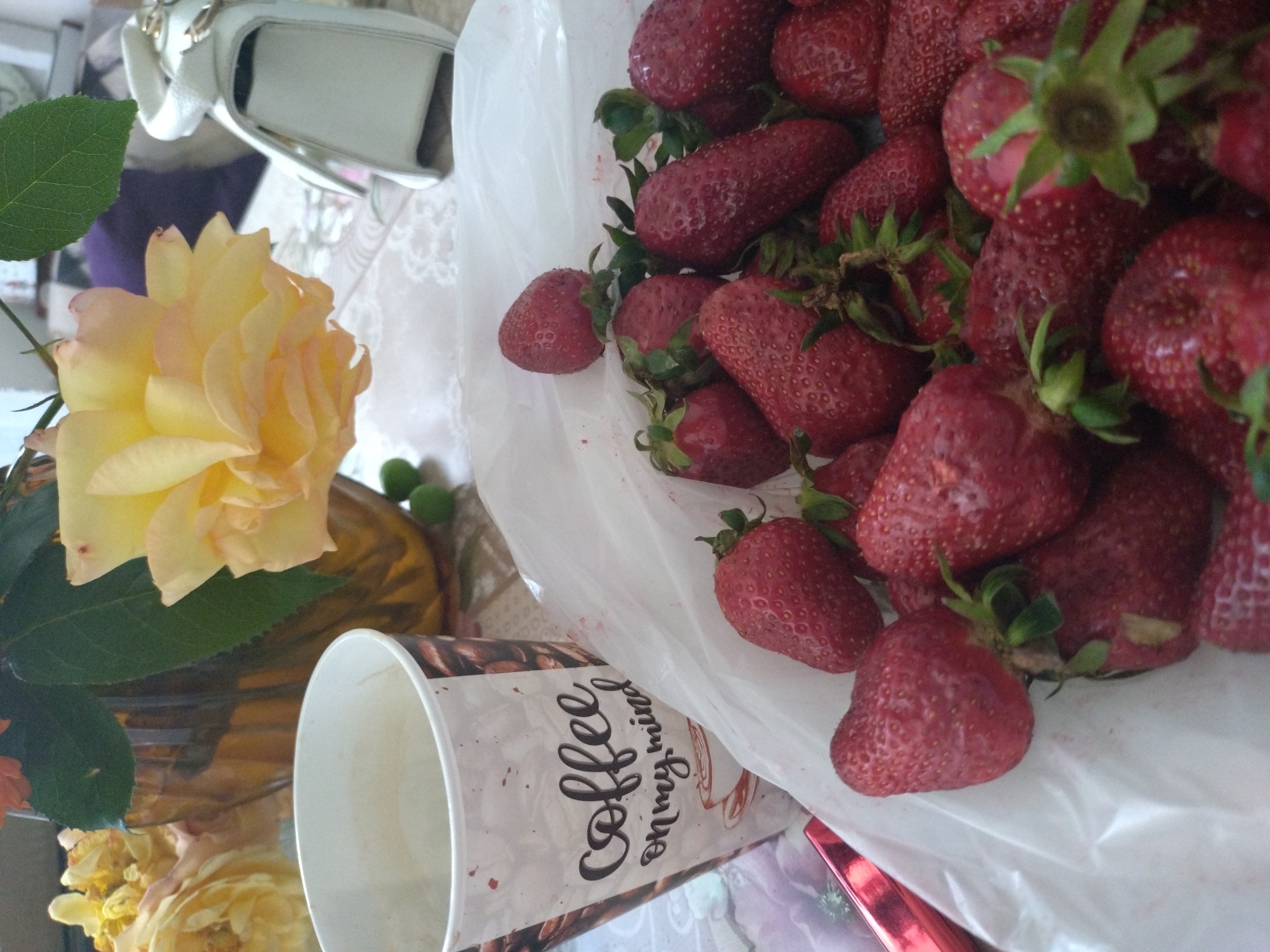 The image shows a still life scene. A plastic bag full of fresh red strawberries is prominently displayed. Next to the bag is a paper coffee cup with the word "coffee" and the phrase "on my mind" printed on it. A vase containing a yellow rose with green leaves is also visible. In the background, parts of a white purse are seen. The items are on a table, likely indoors. There is no indication of the city of Stara Zagora, Bulgaria.