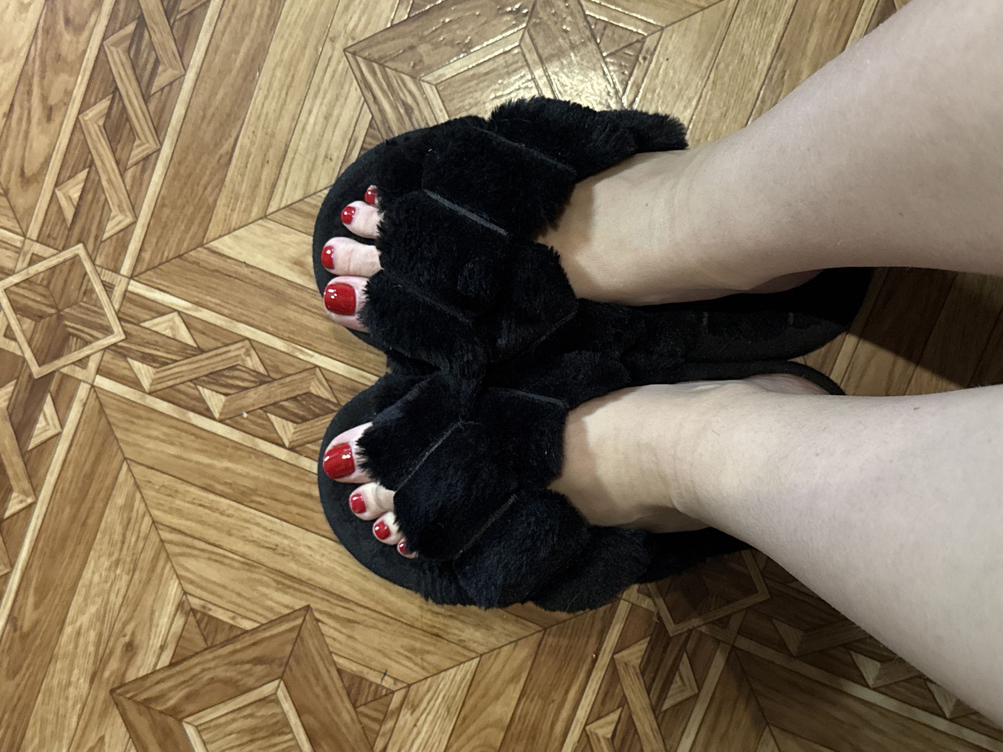 A close-up, overhead shot shows a person's feet wearing black, fluffy slippers with red-painted toenails, positioned on a wooden floor with a geometric pattern. The slippers are open-toed, revealing five toes on each foot with bright red nail polish. The floor is composed of light and dark brown wood planks arranged in a diamond and square pattern. The lighting in the image is diffused, suggesting an indoor environment. No specific location within a city is identifiable.