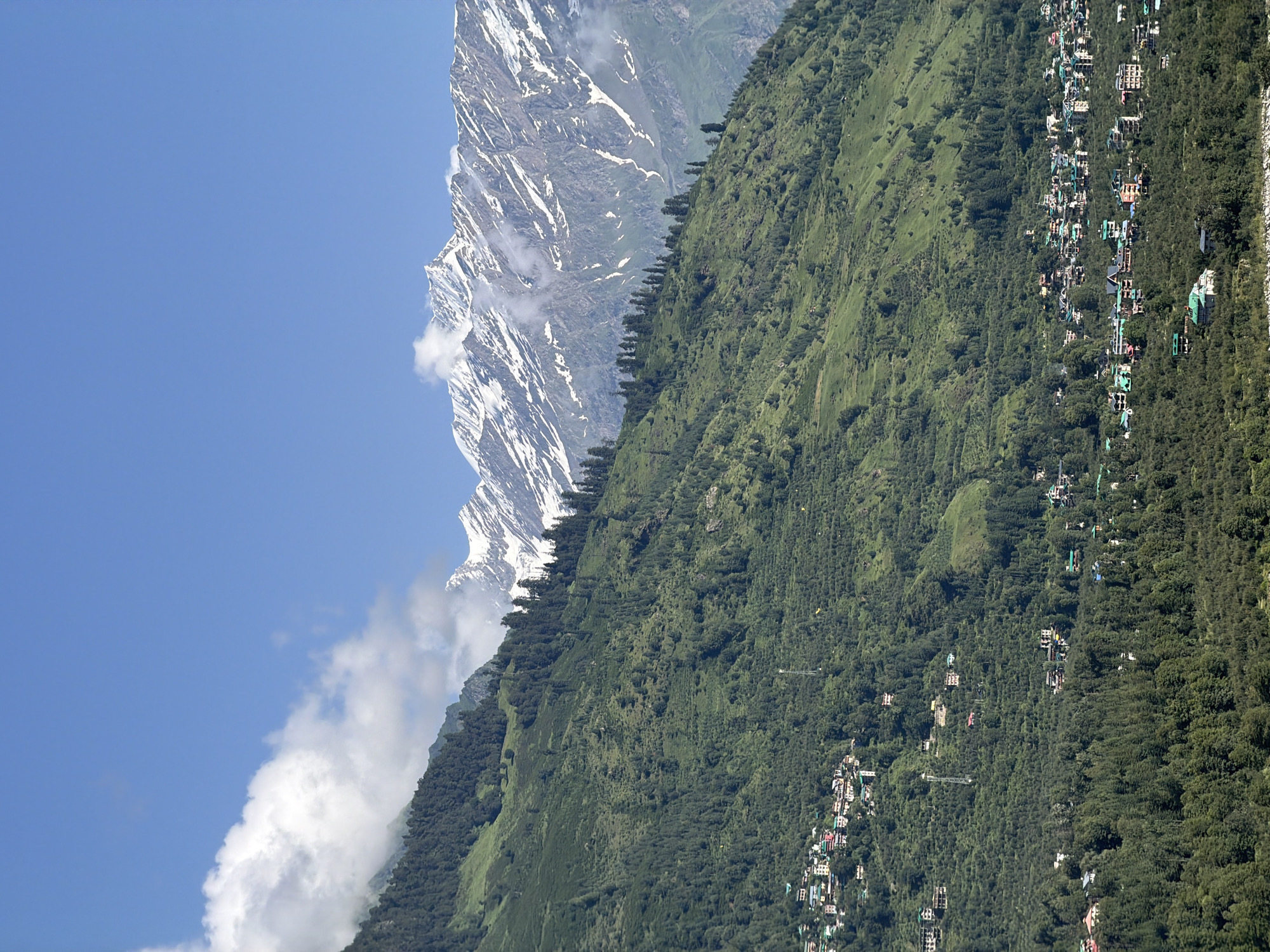 The image shows a panoramic view of the town of Bahang, India, nestled in a lush green valley surrounded by majestic mountains. The foreground is dominated by dense green trees, giving way to terraced fields and scattered houses on the hillside. A few scattered buildings are visible on the lower slopes, gradually becoming denser as they approach the town center. The town itself is a collection of colorful buildings with red and green roofs, clustered together along winding roads. In the background, snow-capped mountains rise majestically against a clear blue sky, with a few wispy clouds adding depth to the scene. The overall impression is one of a tranquil, picturesque mountain village.