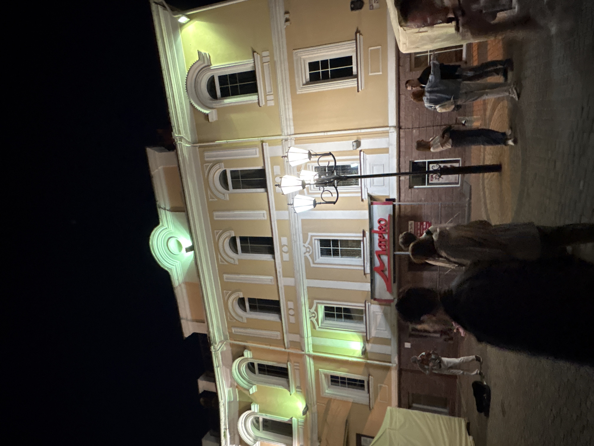 The image depicts an outdoor scene at night, likely in a city. A pale yellow building with arched windows and decorative white trim dominates the background. The building is illuminated with green lights, creating a soft glow. A red sign with the word "Marko" in white letters is visible on the lower facade of the building.
In the foreground and mid-ground, several people are present. On the left, a person dressed in light-colored clothing is playing a violin, with an open bag on the ground in front of them, presumably for donations. Other individuals are standing in small groups or pairs, some facing the building, others interacting with each other. One group appears to be conversing, while another individual is walking away from the camera. A multi-headed lamppost stands near the center of the scene, its lights illuminated.
To the far left, a portion of a light-colored tent or awning is visible. The ground appears to be paved, suggesting a public space like a street or square.
Based on the "Marko" sign, this location is identified as being in the city of Vitsebsk, Belarus, specifically in the vicinity of the Marko building, which houses shops and entertainment venues.