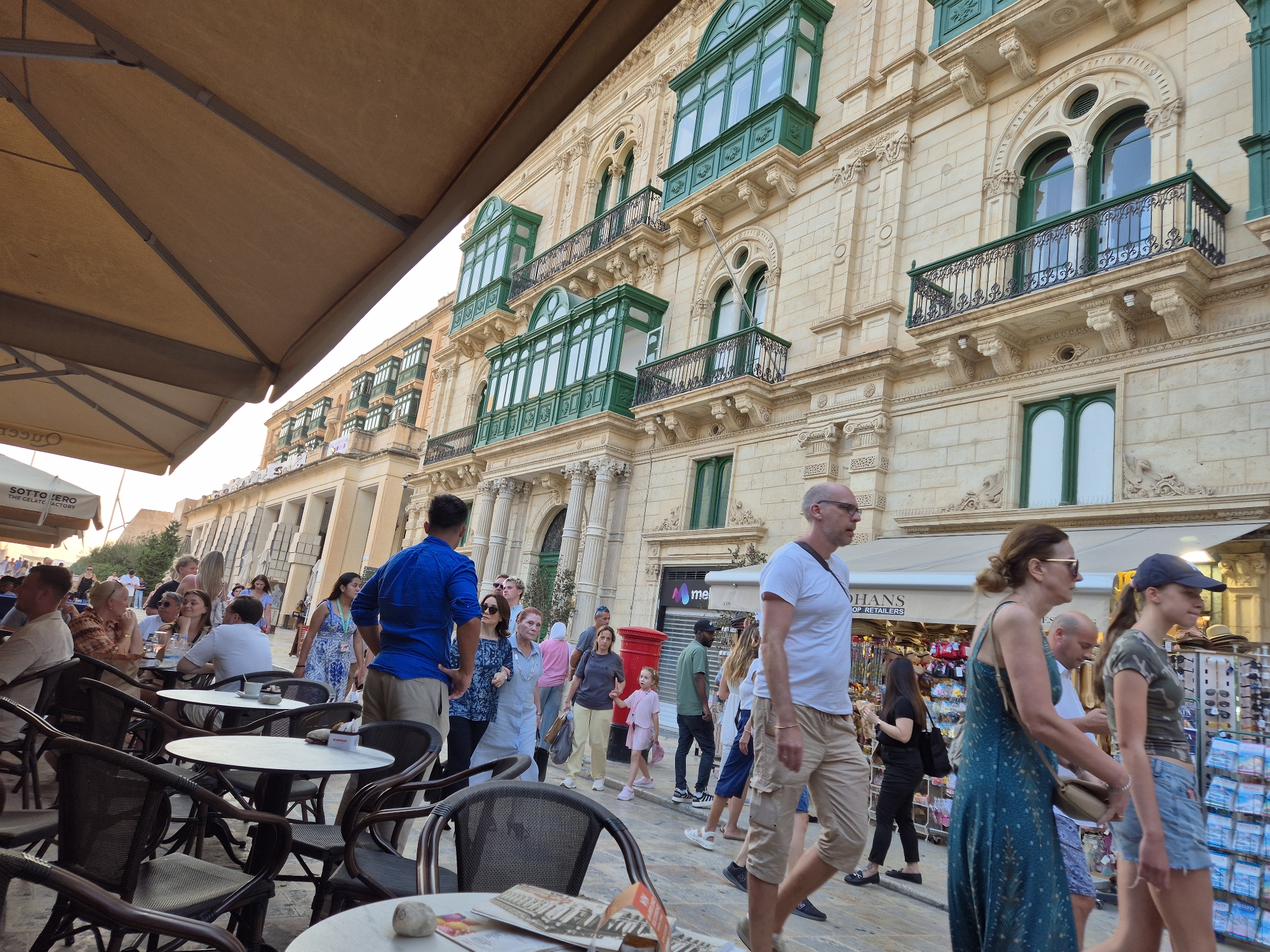 The image depicts a street scene in Valletta, Malta, with a prominent building featuring traditional Maltese balconies. In the foreground, several tables and chairs from an outdoor cafe are visible, some with patrons seated. A large umbrella shades part of the seating area. People are walking on the sidewalk and a street. A man in a blue shirt is standing and looking towards the right. A woman in a patterned blue dress is walking away from the camera. A man in a white t-shirt and cargo shorts is walking towards the camera. Another woman in a green dress is walking towards the camera, and a young woman in a camouflage top and denim shorts is walking ahead of her. The building behind them is a light-colored stone structure with multiple stories, adorned with ornate architectural details and green enclosed balconies, characteristic of Valletta's architecture. The atmosphere appears to be that of a busy tourist area with shops and cafes.