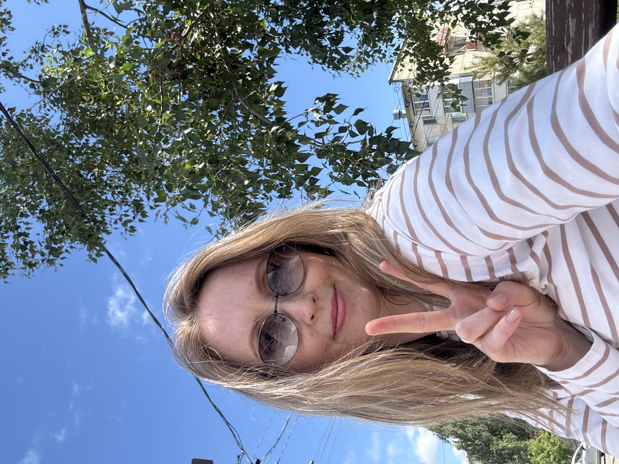 A woman with blonde hair and sunglasses is taking a selfie outdoors. She is wearing a striped shirt and holding up two fingers in a peace sign. The background shows trees, a blue sky, power lines, and a building. The image is taken from a slightly low angle, looking up at the woman. The environment appears to be urban, possibly a park or street in Chaikovsky, Russia, given the architectural style of the building and the presence of deciduous trees common to the region.