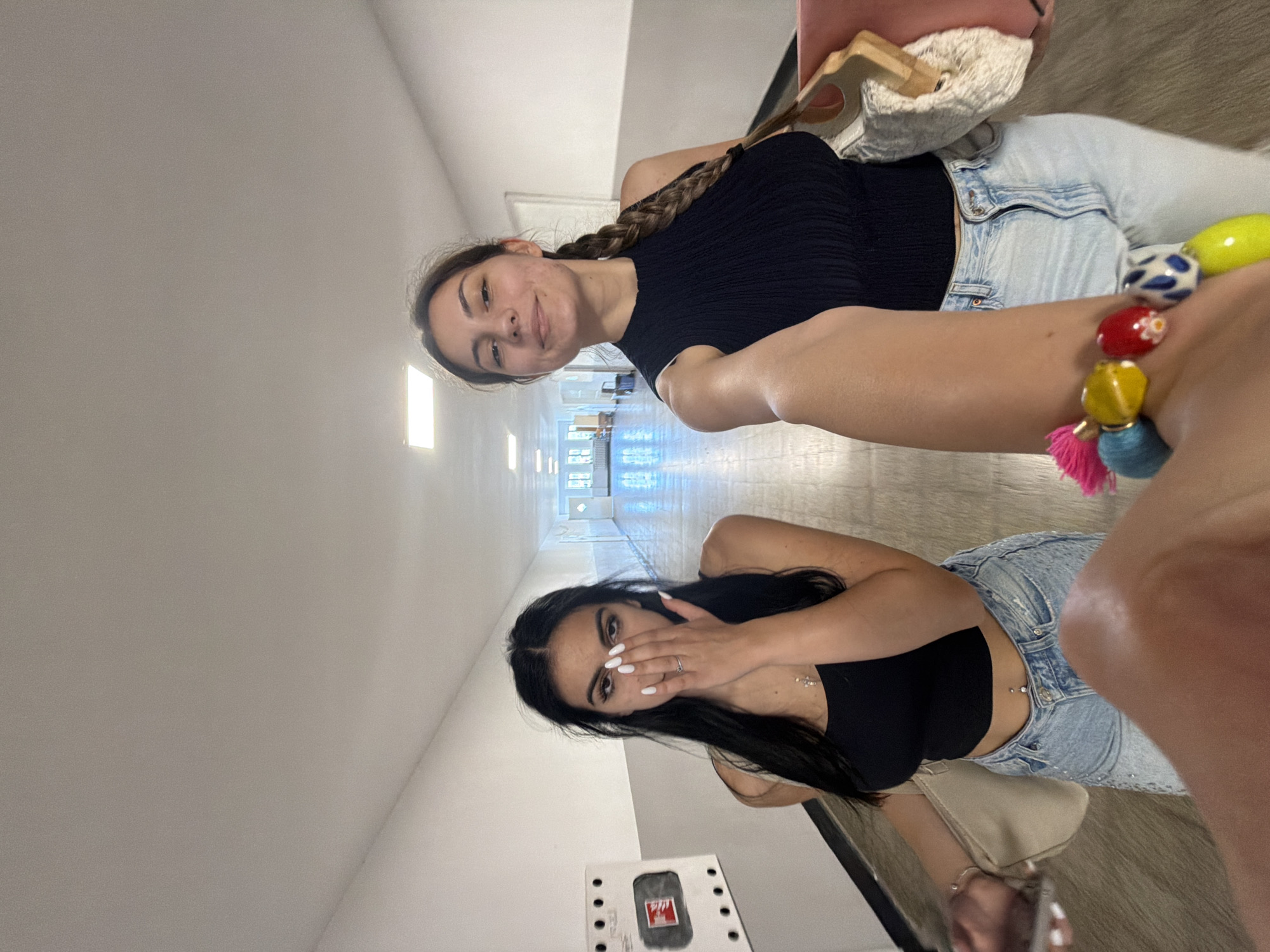 This indoor photograph, taken in Burgas, Bulgaria, features two young women posing in a well-lit hallway. The image is captured from a slightly low, angled perspective.
In the upper part of the frame, a woman with long dark brown hair, styled in a braid, smiles towards the camera. She is dressed in a black ribbed short-sleeved top and light blue high-waisted jeans. Her right arm is visible in the foreground, adorned with a vibrant beaded bracelet featuring red, yellow, blue, and white beads, along with pink and blue tassels.
The lower portion of the frame shows another young woman with long, straight dark hair. She wears a black sleeveless crop top and light blue jeans, with a visible navel piercing. Her hands are raised, covering part of her face in a seemingly playful or shy gesture, showcasing white painted nails and rings on her fingers, including a silver-colored ring on her left ring finger. A small, beige shoulder bag is visible by her side.
The setting is a long, bright corridor or hallway, characterized by light-colored walls and a reflective, light-toned floor, possibly made of tiles or polished concrete. Fluorescent light fixtures are visible on the ceiling, providing illumination. The hallway extends into the background, revealing additional lighting and what appear to be windows or openings on the right side. On a nearby wall, a white rectangular panel with a red symbol inside a black square is mounted, though no specific text is clearly legible. The overall atmosphere is casual and relaxed.