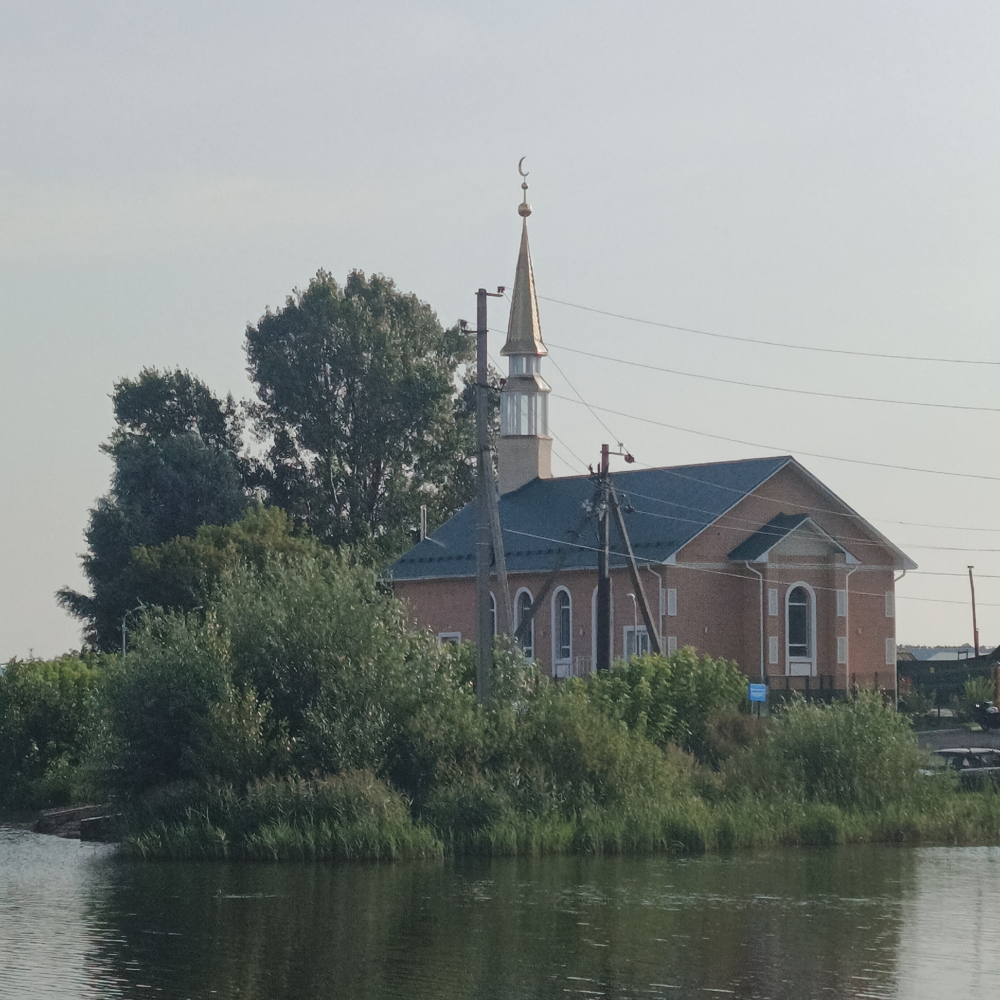 The image shows a mosque situated on the bank of a body of water, likely a river or lake, in the city of Kambarka, Russia. The mosque is a two-story brick building with a green roof and a tall, slender golden minaret topped with a crescent moon. The building has arched windows and decorative white elements around them. Lush green trees and vegetation surround the mosque and extend to the water's edge. Several utility poles with wires are visible in the foreground and to the sides of the mosque. The sky is overcast and pale. The water in the foreground is dark and reflects the surrounding greenery. There are no people visible in the image.