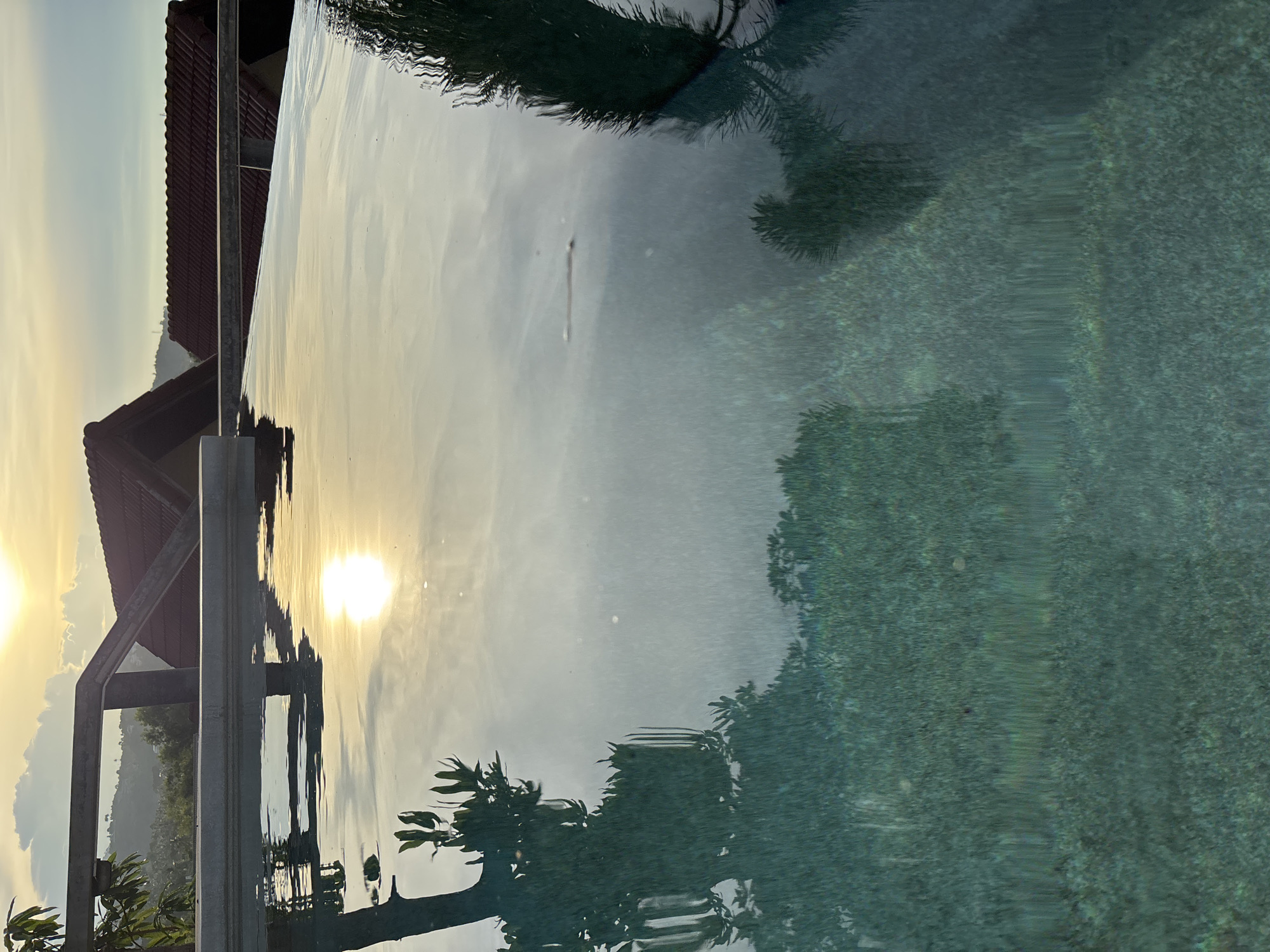 The media file shows a reflective surface, likely a swimming pool, with a clear view of the sky and surrounding landscape. The sun is visible, casting a bright glow and creating reflections on the water. The time of day appears to be either sunrise or sunset, due to the low angle of the sun and the warm, golden light. The sky is partly cloudy, with soft hues of yellow and blue.
In the background, there is a building with a reddish-brown tiled roof, suggesting a tropical or subtropical location. The building seems to be part of a resort or residential property, possibly a house given the "บ้านบางรักษ์, Thailand" location context. A metal railing is also visible, extending along the edge of the reflective surface.
The water's surface acts as a mirror, reflecting the sky, the sun, and the silhouettes of trees. The reflections are distorted by ripples on the water, adding a dynamic quality to the scene. The overall mood is peaceful and serene, with a sense of tranquility. The image captures a moment of natural beauty and calm. No text is visible in the media file.
