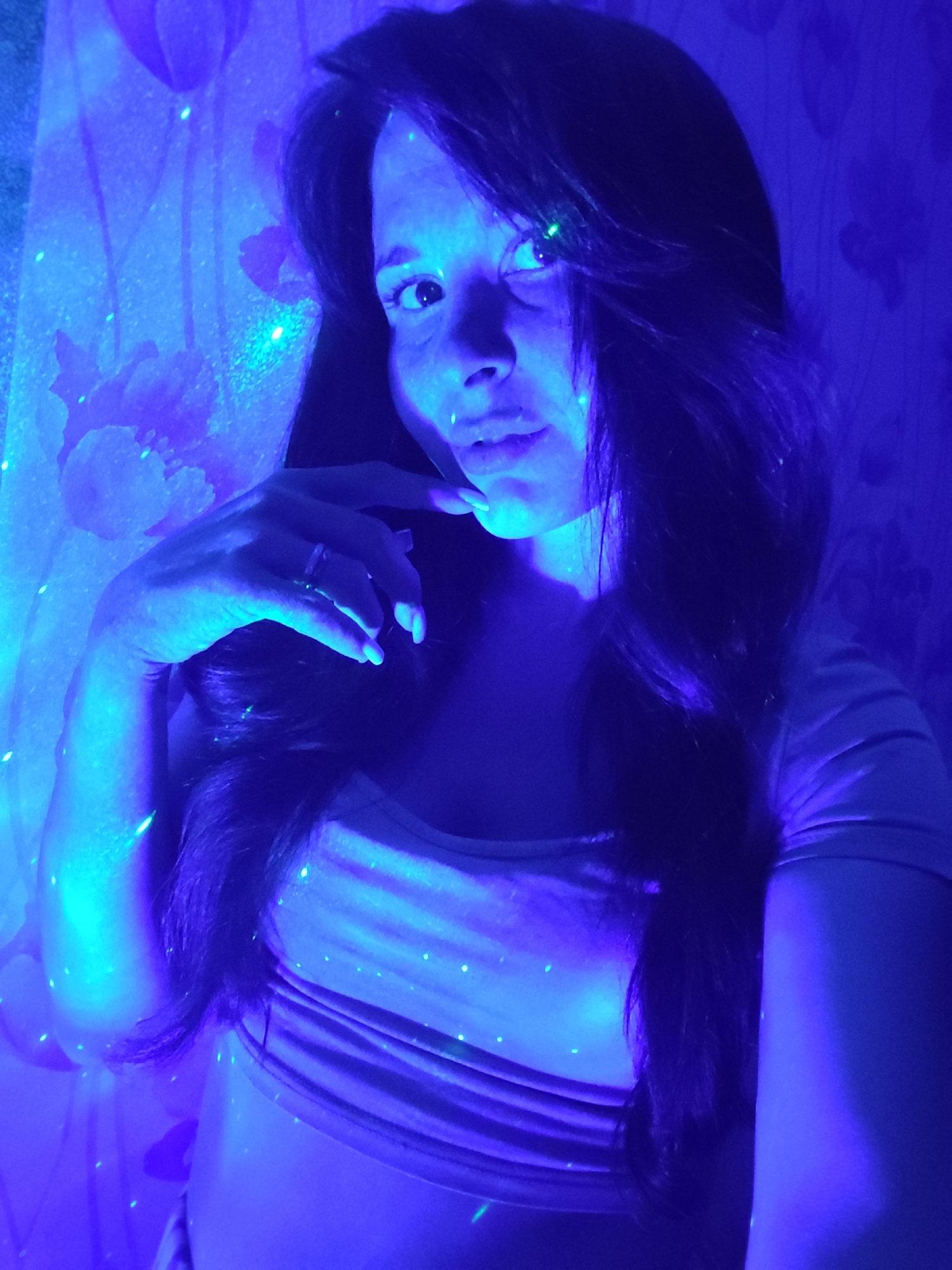 A woman poses in a room illuminated by blue light, possibly from a projector or light source, as evidenced by the scattered points of light on her skin, hair, and clothing. She is wearing a light-colored top and a ring is visible on her finger. The background includes a wall with a floral pattern. The presence of the city of Фоки, Russia is uncertain in this image.