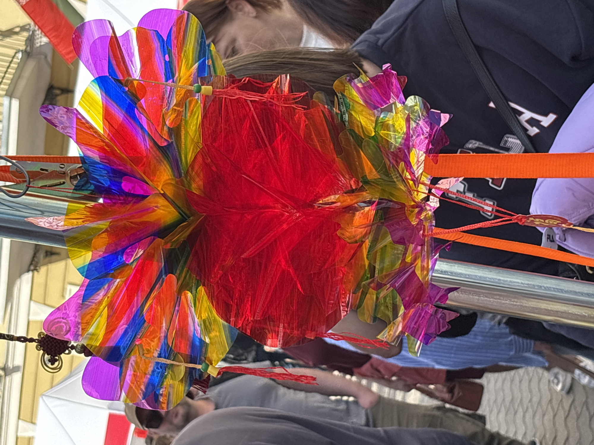 The image shows a colorful, decorative object resembling a flower or pinwheel, made of translucent, crinkled plastic in various vibrant hues like red, yellow, blue, purple, and orange. This object is suspended from a pole and appears to be at an outdoor market or street fair. In the background, several people are visible, blurred due to shallow depth of field. One person in the foreground is wearing a dark-colored hooded sweatshirt with a white logo. The ground is paved with cobblestones. The scene suggests a lively public gathering. The context of Minsk, Belarus, implies this could be a local festival or event, though specific identifying landmarks are not present in the image.