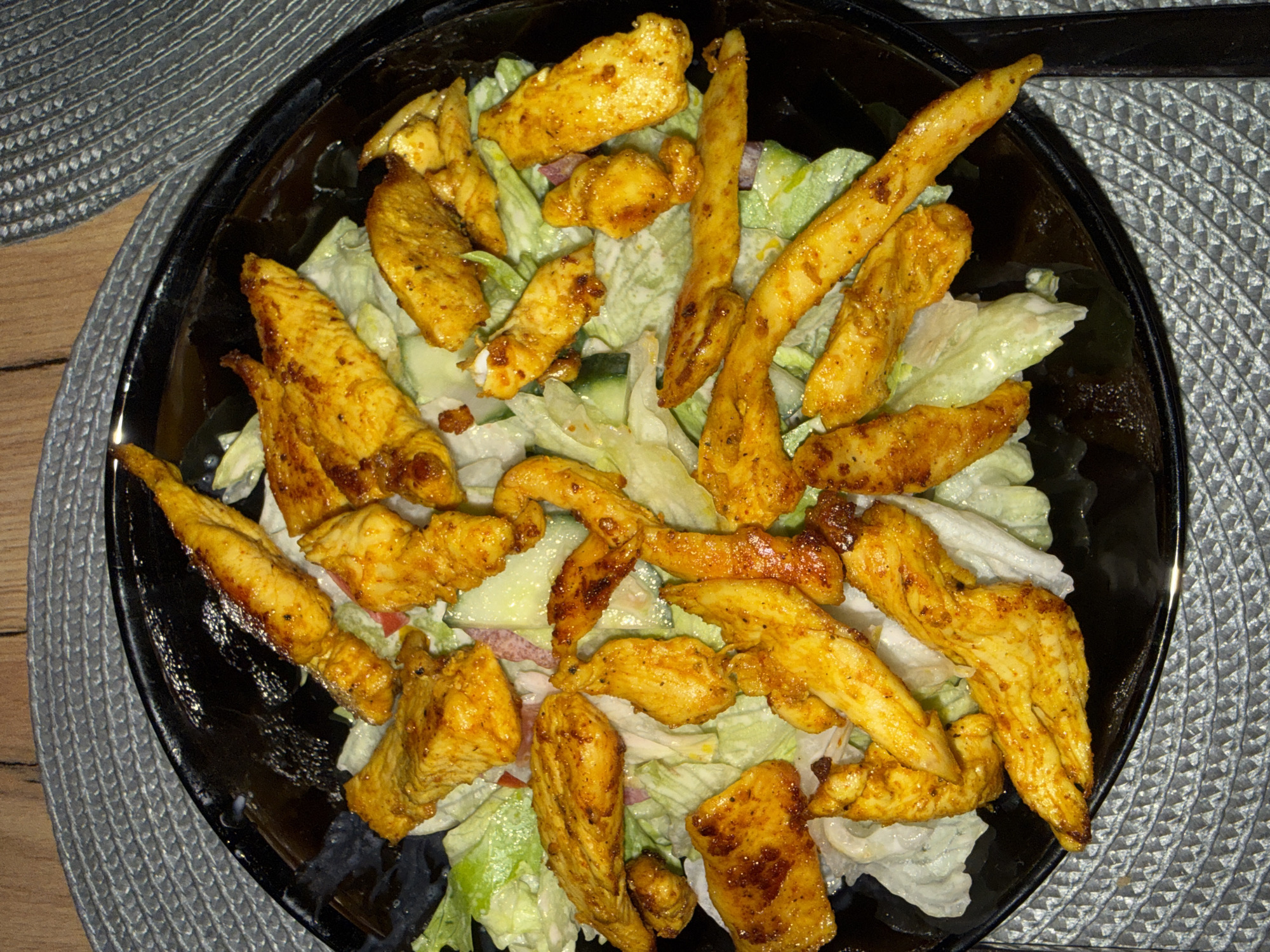 A close-up, overhead shot shows a black, circular plate filled with a colorful salad. The salad consists of shredded iceberg lettuce, sliced cucumbers, and what appears to be thin slices of red onion. The main focus of the salad is numerous pieces of golden-yellow, seasoned, grilled chicken strips. The chicken pieces are coated in spices, giving them a slightly charred and well-cooked appearance. The plate is positioned on a textured gray placemat, which sits on a wooden table. The lighting is somewhat dim, with the focus primarily on the food. There is no indication of the location being in Varna, Bulgaria or any other specific location.