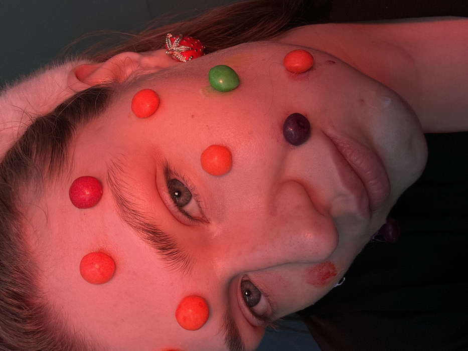 The image displays a close-up, high-angle portrait of a human subject's face, likely female, illuminated predominantly by a red-pink light source originating from the left. The subject has light-colored irises and dark, defined eyebrows. A light-colored, fuzzy headband is visible across the subject's hairline. The subject's right ear is adorned with a red, textured earring shaped like a strawberry. Approximately fifteen spherical, candy-like objects, primarily red, are adhered to the facial skin, distributed across the forehead, temples, cheeks, and chin. Specifically, one green spherical object is located on the right cheek, and two dark purple spherical objects are present on the left cheek and below the left corner of the mouth. A third dark purple object is partially visible hanging below the chin on the left side. Reddish smudges, one circular with a raw appearance, are observed on the left cheek inferior to the left eye. A small, clear droplet or bead is visible on the subject's right jawline. The subject wears a dark-colored garment, visible on the lower left of the frame. The background is an out-of-focus, dark interior, exhibiting a greenish-blue hue in areas under the ambient lighting. The subject maintains direct eye contact with the viewer. The location of the scene is Kazan, Russia.