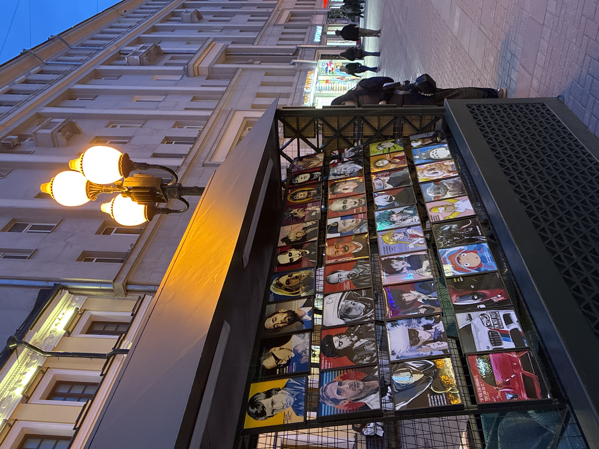 A vertical, eye-level shot captures a street scene in Moscow, Russia, at dusk. A multi-story building with rows of windows and architectural details fills the left and upper background. A vintage streetlight with globe-shaped bulbs illuminates the immediate foreground.
To the right of the streetlight, a metal structure displaying a collection of graphic art pieces stands out. The art appears to be mounted on panels within the structure's frame.
Further along the street, figures of pedestrians walking on the sidewalk can be seen. A brightly lit store with signage in Cyrillic script is also visible. The overall scene is a mix of urban architecture, street lighting, and pedestrian activity, indicative of a city environment.