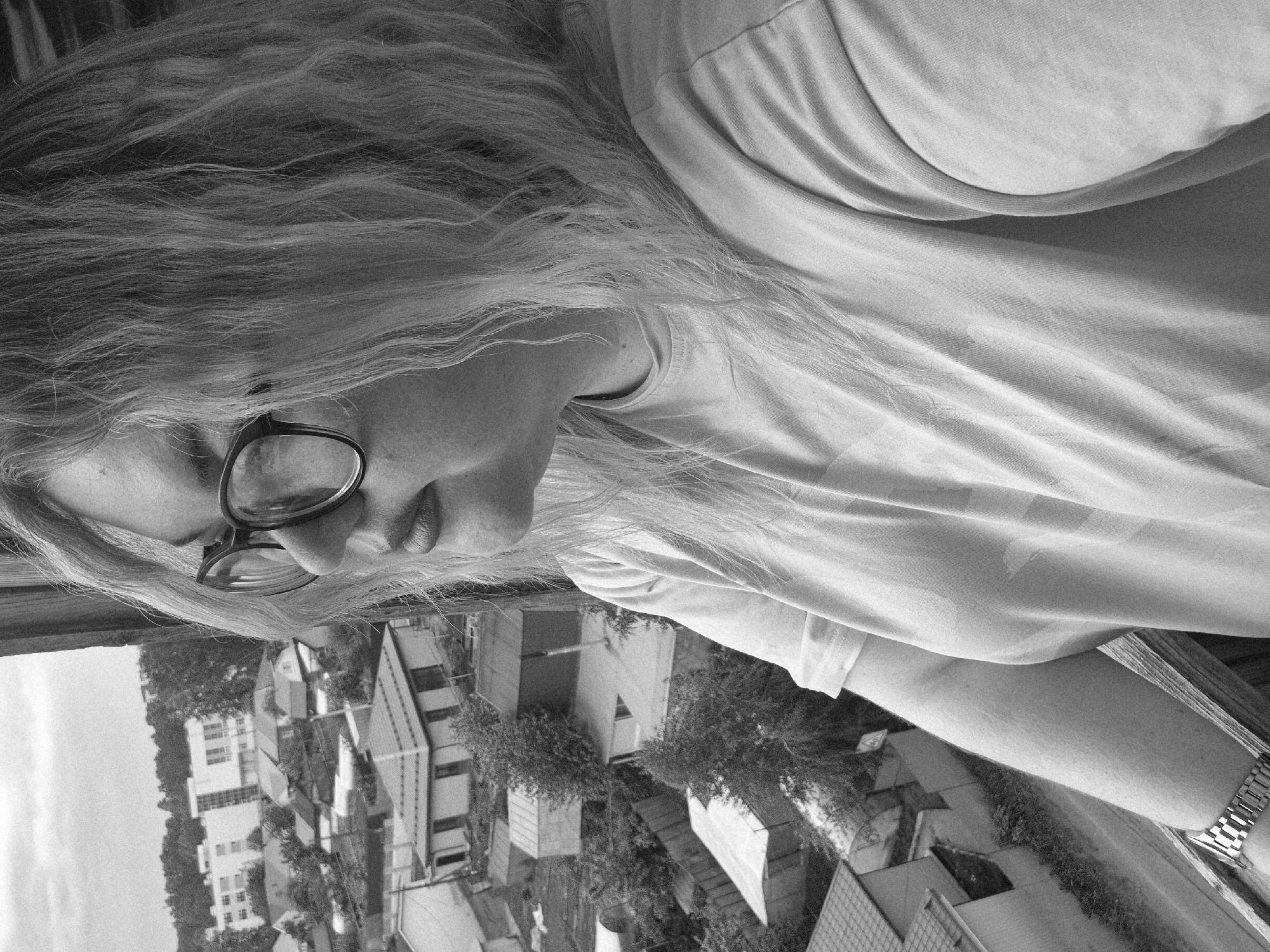 A grayscale, eye-level shot of a person wearing glasses, looking down towards a cityscape. The person has long hair and is wearing a t-shirt and a watch on their wrist. The background features buildings and trees, characteristic of an urban area. The city is Chaikovsky, Russia.