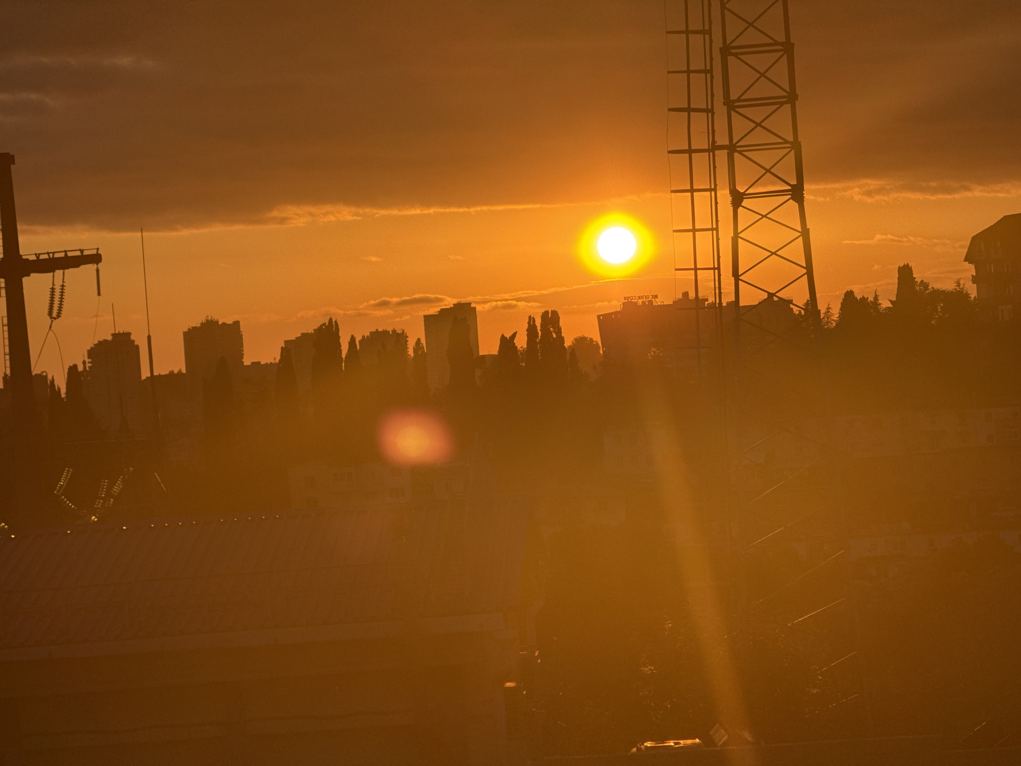 This media file captures a vibrant sunset over a city skyline, likely in Sochi, Russia. The dominant subject is the sun, a brilliant orb of yellow-white light, positioned in the upper center of the frame. It casts a warm, golden-orange glow across the entire scene, creating a hazy and atmospheric effect.
The skyline is composed of silhouetted buildings of varying heights, interspersed with dark, conical shapes that suggest pine trees, typical of coastal or mountainous regions like Sochi. In the foreground, the roof of a building with a slightly metallic sheen is visible, dotted with what appear to be small lights. There is also a large, dark, lattice-like structure, resembling a tower or part of a crane, on the right side of the frame, and another similar structure on the far left, partially obscured.
The sky is a blend of deep orange and darker hues, with faint, wispy clouds catching the sunset's light. The overall impression is one of a peaceful end to the day, with the intense sunlight creating lens flares and light rays that add to the visual drama. The time of day is clearly sunset, and the weather appears clear, with the sun being the primary source of illumination. The image evokes a sense of warmth and tranquility. There is some faint text visible on a building in the middle ground, but it is too blurred to be legible.