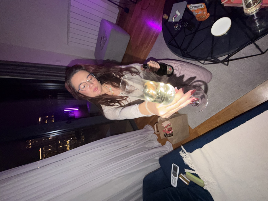 The image displays an indoor scene at night, featuring a female subject within what appears to be a modern apartment in Belgrade, Serbia. Through a large window or balcony door, a nighttime cityscape with illuminated high-rise buildings is visible, suggesting an urban residential area such as Belgrade Waterfront or Novi Beograd.
The subject is a female with long, dark brown hair and glasses, wearing a light-colored, long-sleeved top and striped pants. She is kneeling or crouching on a grey carpeted floor. Her right hand, adorned with dark red nail polish, extends a clear champagne flute containing a sparkling pale yellow liquid directly towards the viewer. In her left hand, she holds a green bottle labeled "Martini Asti." Her facial expression shows pursed lips and her gaze is directed slightly downward.
The immediate environment includes a dark blue sofa or armchair partially covered by a white textured throw blanket on the left, with a white remote control and a mobile phone resting on its armrest. A brown paper bag with red handles sits on the wooden floor between the sofa and the subject. To the right, a dark, glass-topped coffee table with a geometric metal base holds a white coaster, a small silver container, another pair of glasses, and an orange snack bag. In the background, against a light-colored wall, a grey ottoman or footstool is visible next to a white wall-mounted radiator. Purple ambient light emanates from a vertical fixture near the window. White sheer curtains are drawn back on the left, revealing the exterior night scene. The flooring consists of wooden planks near the window and sofa, transitioning to grey carpet in the central area.