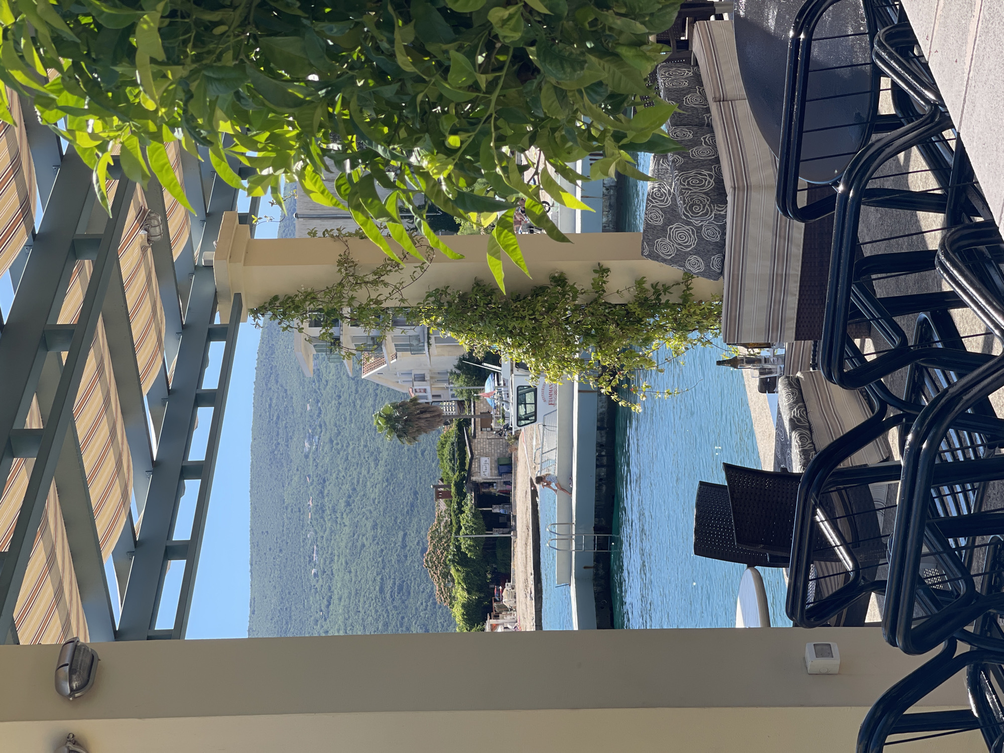 The image depicts an outdoor dining or lounge area overlooking a bay or coastal inlet in Đenovići, Montenegro. In the immediate foreground, a pergola with a teal-colored metal frame supports striped fabric awnings, providing shade. A silver-colored marine-style bulkhead light fixture is attached to a light-colored structural column on the left. In the lower foreground, multiple black metal chairs with slat designs are arranged, some appearing stacked. Behind these chairs, a wicker-style outdoor sofa with striped upholstery and gray cushions featuring a white floral pattern is visible. A small glass table with a glass and a brown bottle is positioned next to the sofa. A light-colored column covered in climbing green vines stands centrally, with another light fixture partially visible above. A small white electrical box is mounted on the left column.
The midground features calm, clear blue-green water. A concrete pier extends from the right, equipped with a metal ladder descending into the water. A person, appearing to be a child or small adult, is seated on the edge of the pier, facing away from the viewer, with their feet positioned above the water. A white boat named "FLAMMAN" with "RESTAURANT" printed above its name, is docked to the right of the person. Beyond the pier, a narrow shoreline is visible with a small beach area, modest buildings, and dense foliage, including a prominent palm tree. One building on the shore has "Marija" written on it.
The background is dominated by a range of densely forested mountains under a clear, bright blue sky. Scattered white residential buildings with red-tiled roofs are visible on the lower slopes of these mountains and along the distant coastline. A large bush with vibrant green leaves partially obscures the right side of the midground and background.