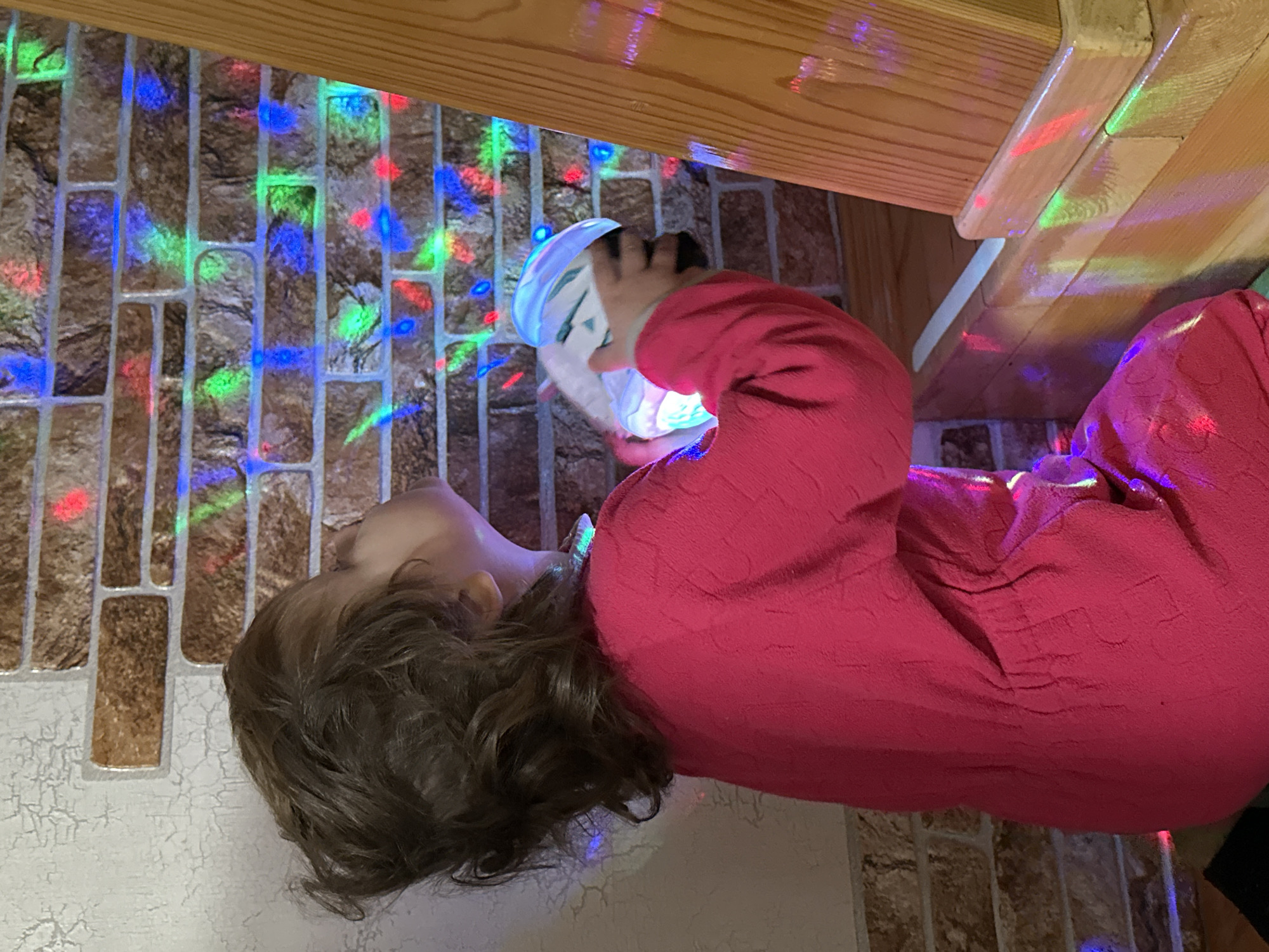 A young child, wearing a pink, long-sleeved garment, is holding a lit-up toy object with both hands. The child's head is tilted back, and their brown hair is visible. The child is positioned near a wall adorned with brick-like tiles illuminated by multicolored lights. Overhanging the scene is a section of a wooden structure. The location is possibly in an interior setting in Chaikovsky, Russia.