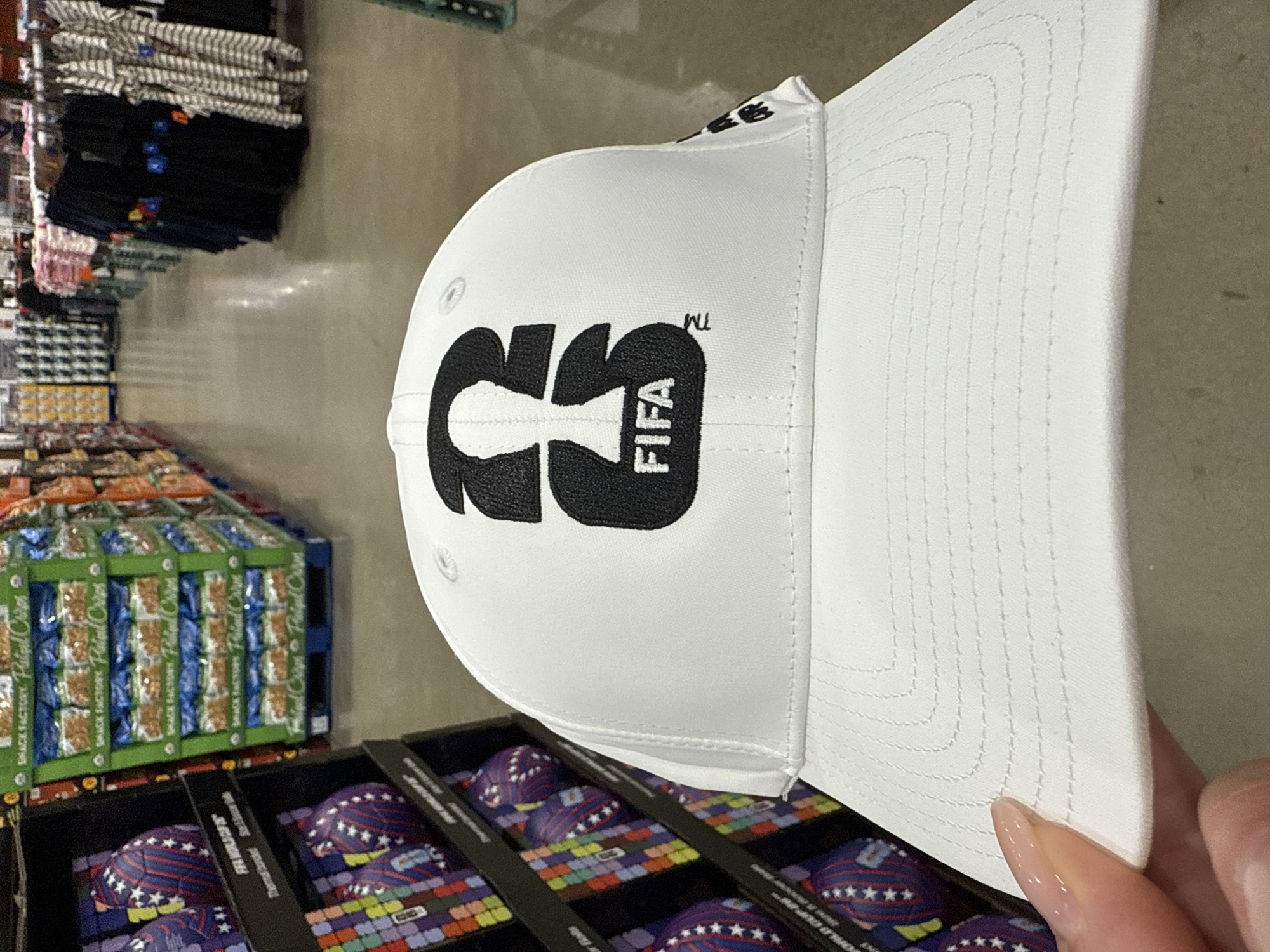 The image shows a white FIFA-branded baseball cap resting on top of a display case. The cap has the FIFA logo embroidered in black. The display case contains soccer balls adorned with a multi-colored pattern and white stars. The balls appear to be packaged for retail sale. Behind the display case is a pallet loaded with boxes of Snack Factory Pretzel Crisps. In the background, there is a metal shelf unit with various stacks of clothing items. The scene appears to be in a warehouse store setting, potentially in Bolingbrook, United States of America. A fingernail belonging to a person is visible in the bottom right corner of the image.
