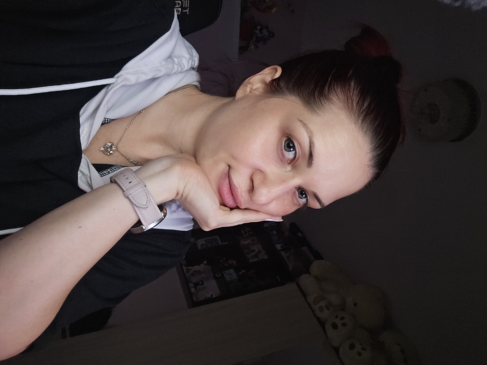 The image portrays a young woman, likely in her 20s or 30s, in an indoor setting. She is positioned at an angle, resting her head gently on her left hand, and looking directly towards the camera with a calm expression and a subtle smile.
She has dark hair tied back in a bun and striking light-colored eyes. She is dressed in a black and white hooded top or sweatshirt. A delicate gold chain with a small, intricate pendant adorns her neck. On her left wrist, a light pink smartwatch or fitness tracker with a silver clasp is visible. Faint white text, reading "ET," can be seen on the black fabric of her top near her left shoulder.
The background is dimly lit and largely out of focus, suggesting low light conditions, possibly evening or nighttime. Visible elements include a blurry circular object, potentially a light fixture, in the upper right. In the lower right, a cluster of light-colored, soft toys or plushies is discernable. Behind her head, a dark surface with faint patterns hints at an indoor environment. The overall mood conveyed is one of relaxation and quiet contemplation. The known location for this media file is Burgas, Bulgaria.