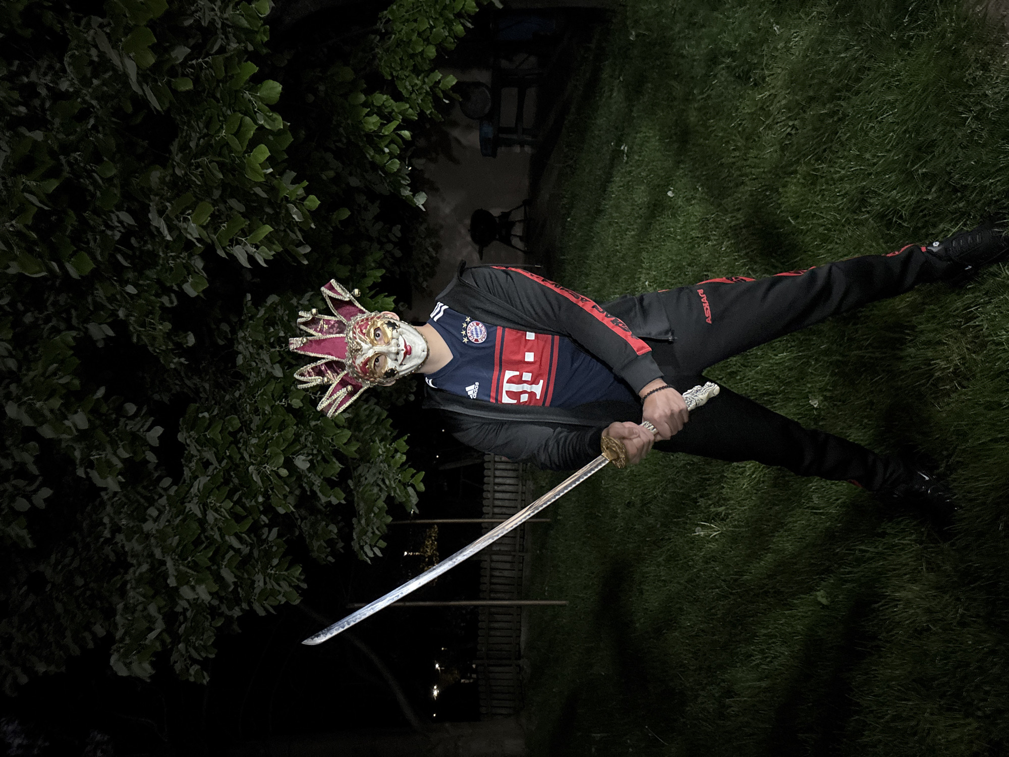 A person wearing a red and gold Venetian jester mask, a dark blue Bayern Munich soccer jersey under a dark jacket with red stripes on the sleeves, and dark pants and shoes, holds a katana with a silver blade. The person stands on a patch of dark green grass, with a tree on the left side of the frame. In the background, a dark pathway leads into the distance, with what appears to be a small structure in the background. The scene is dimly lit, suggesting it is nighttime. There is no information regarding the location in the city of Goritsa, Bulgaria.