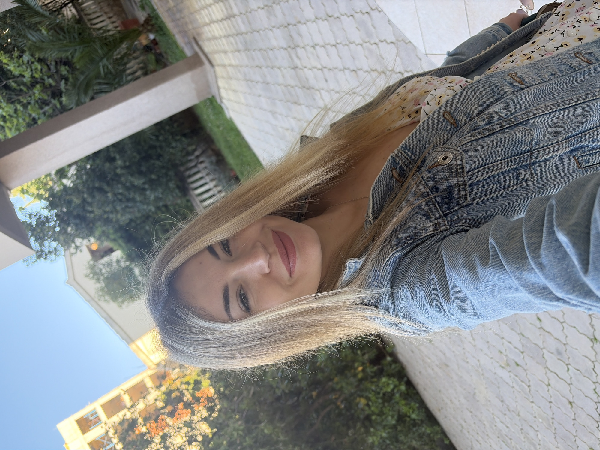 A medium shot depicts a woman with blonde hair, wearing a denim jacket over a floral dress, located in Budva, Montenegro. The backdrop includes a view of a building with a beige facade and multiple windows, trees, and a paved pathway.