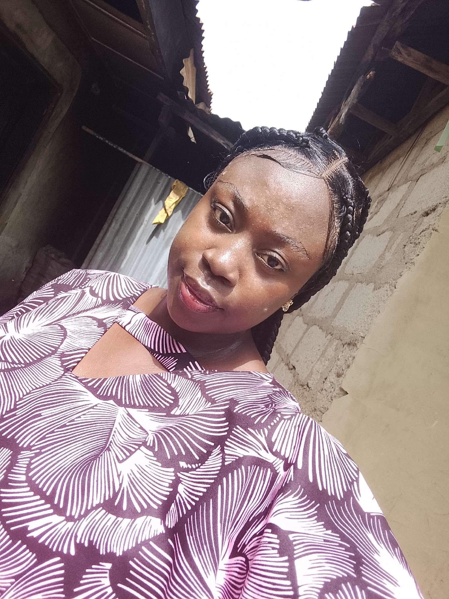 A young woman poses for a selfie in Akwanga, Nigeria. She wears a brown and white patterned top with a small triangular cutout at the collarbone. Her hair is styled in braids. The background includes a building wall, corrugated metal sheeting, and a partial view of the roof.