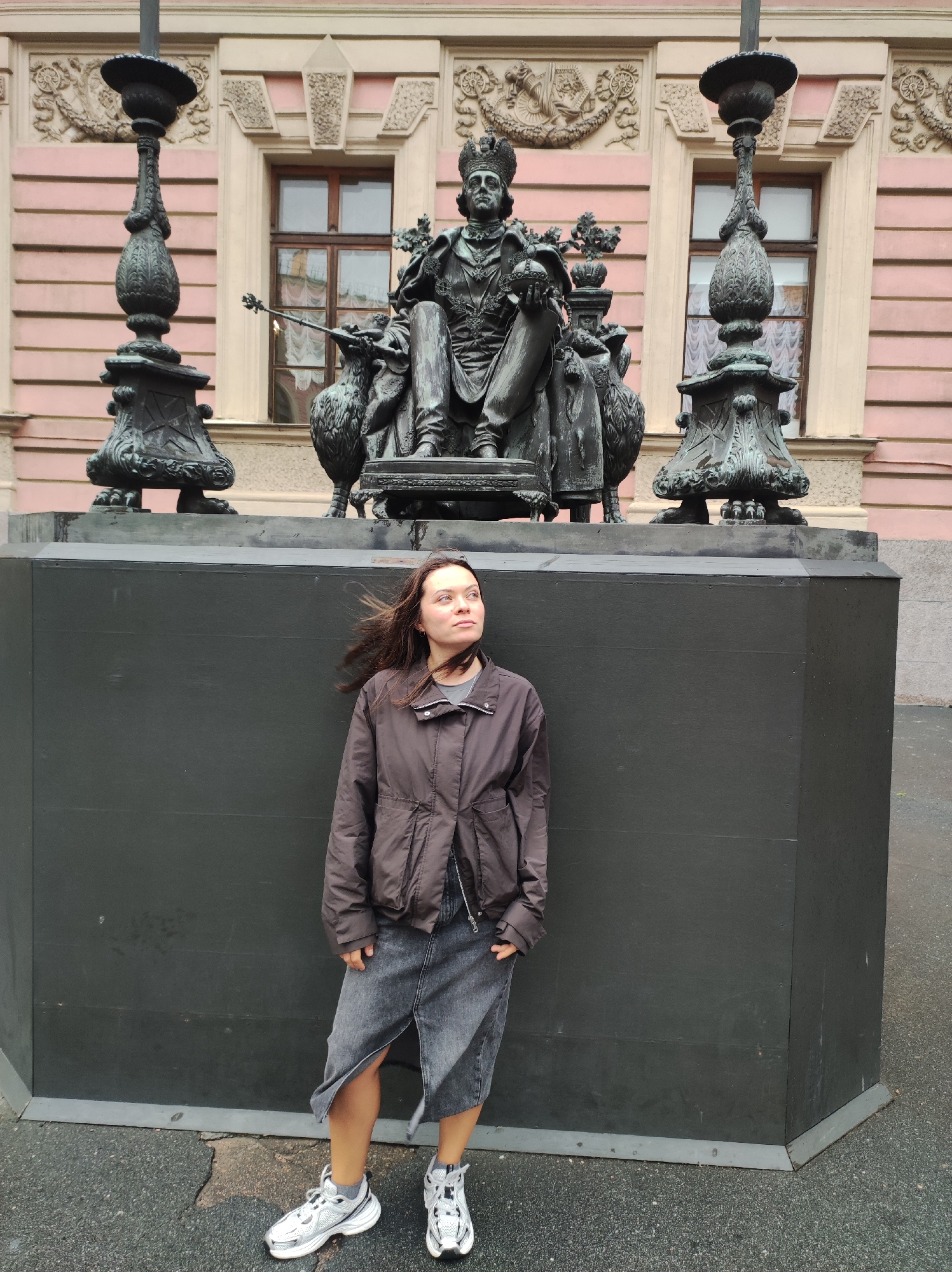 A young woman stands in front of a dark gray, rectangular pedestal in front of a statue and building in Saint Petersburg, Russia. The woman is wearing a dark brown jacket, a dark gray denim skirt, and white and gray sneakers. She is looking to her right, with her hair blowing in the wind. The statue behind her depicts a seated figure wearing a crown and robes, holding a scepter and orb. The statue is bronze and rests on a dark pedestal. Behind the statue is a light pink building with arched windows. The ground is paved with asphalt.