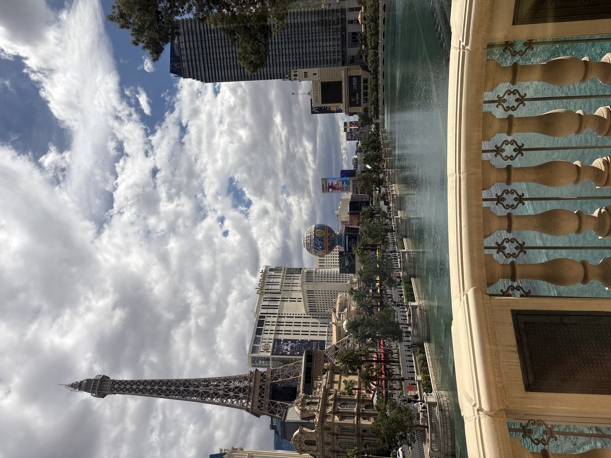 The image is taken from a high vantage point, likely a balcony, overlooking the Las Vegas Strip in Paradise, Nevada, United States. A decorative balustrade runs along the right edge of the frame. In the foreground, a water feature or lake reflects the sky and cityscape. The Paris Las Vegas Hotel and Casino with its replica Eiffel Tower is prominent. The Balloon of the Paris Hotel is seen along with other casinos, hotels, billboards, and buildings. The sky is filled with white and grey clouds.